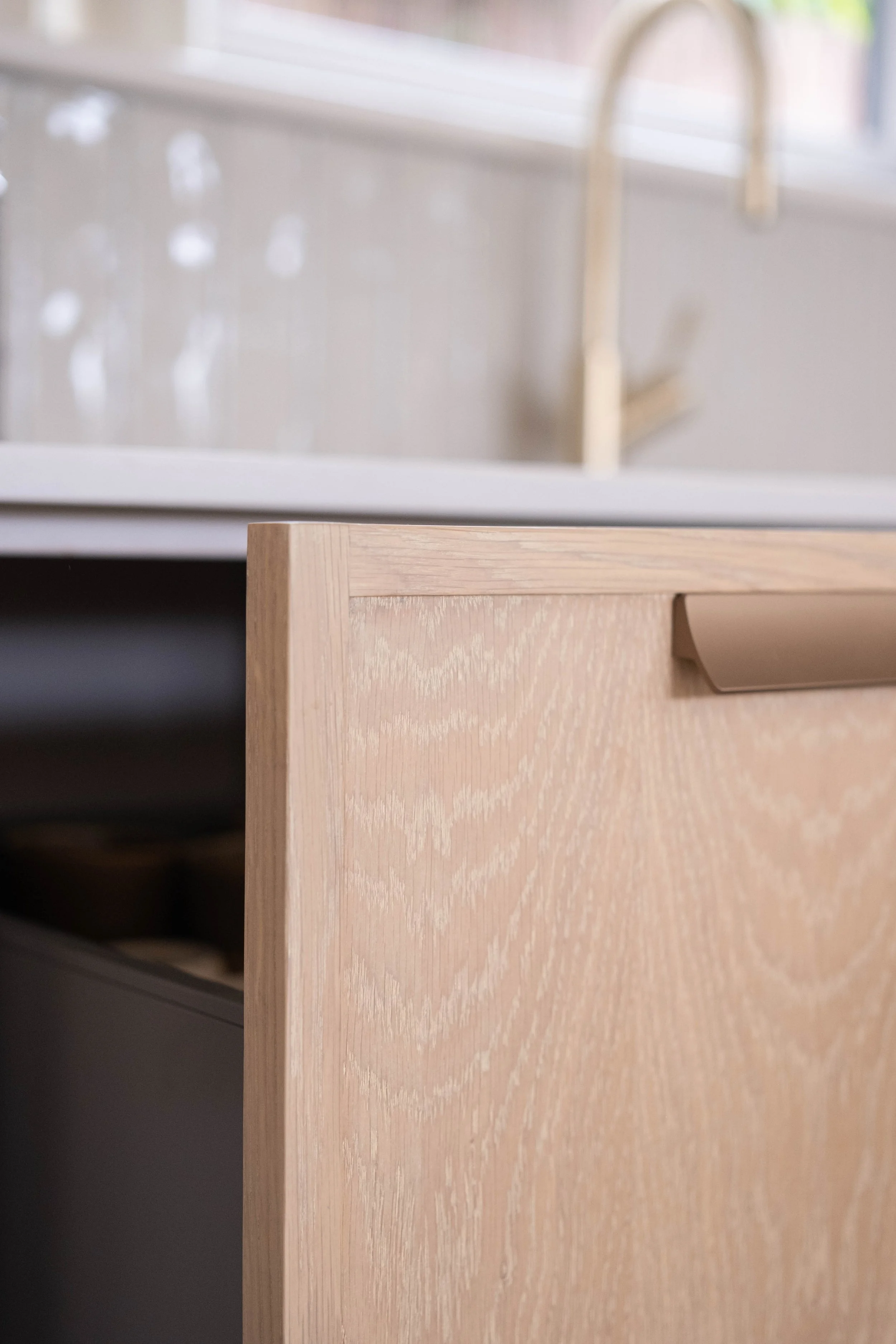Close-up of a light wood-colored drawer with a rectangular handle, part of a wooden furniture piece, with a white kitchen counter and a gold-colored sink faucet in the background.