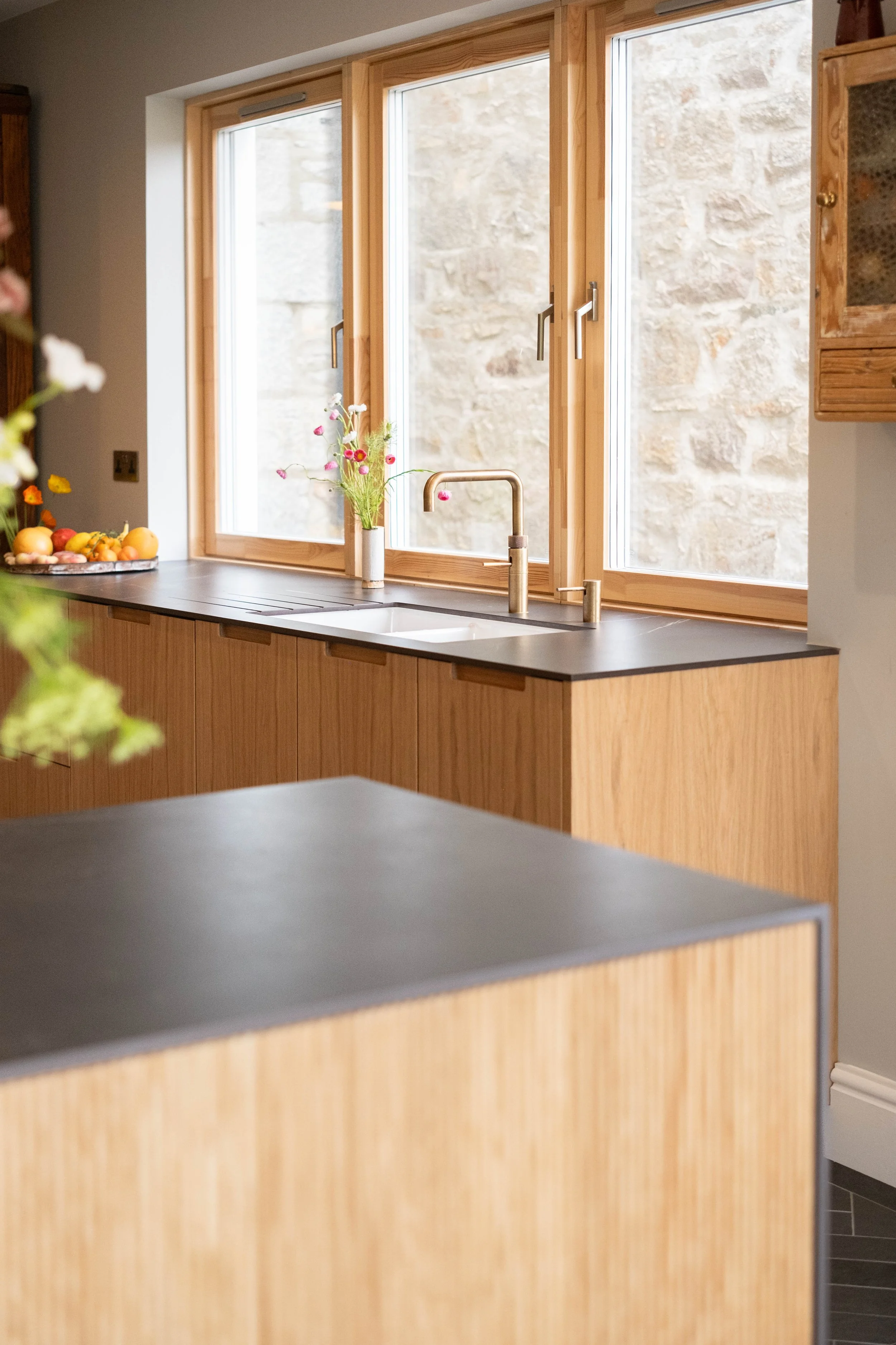 A kitchen with three large sliding windows above a wooden counter with a black surface, a vase of pink and white flowers, and a bowl of assorted fruits. The background outside the windows is a stone wall.