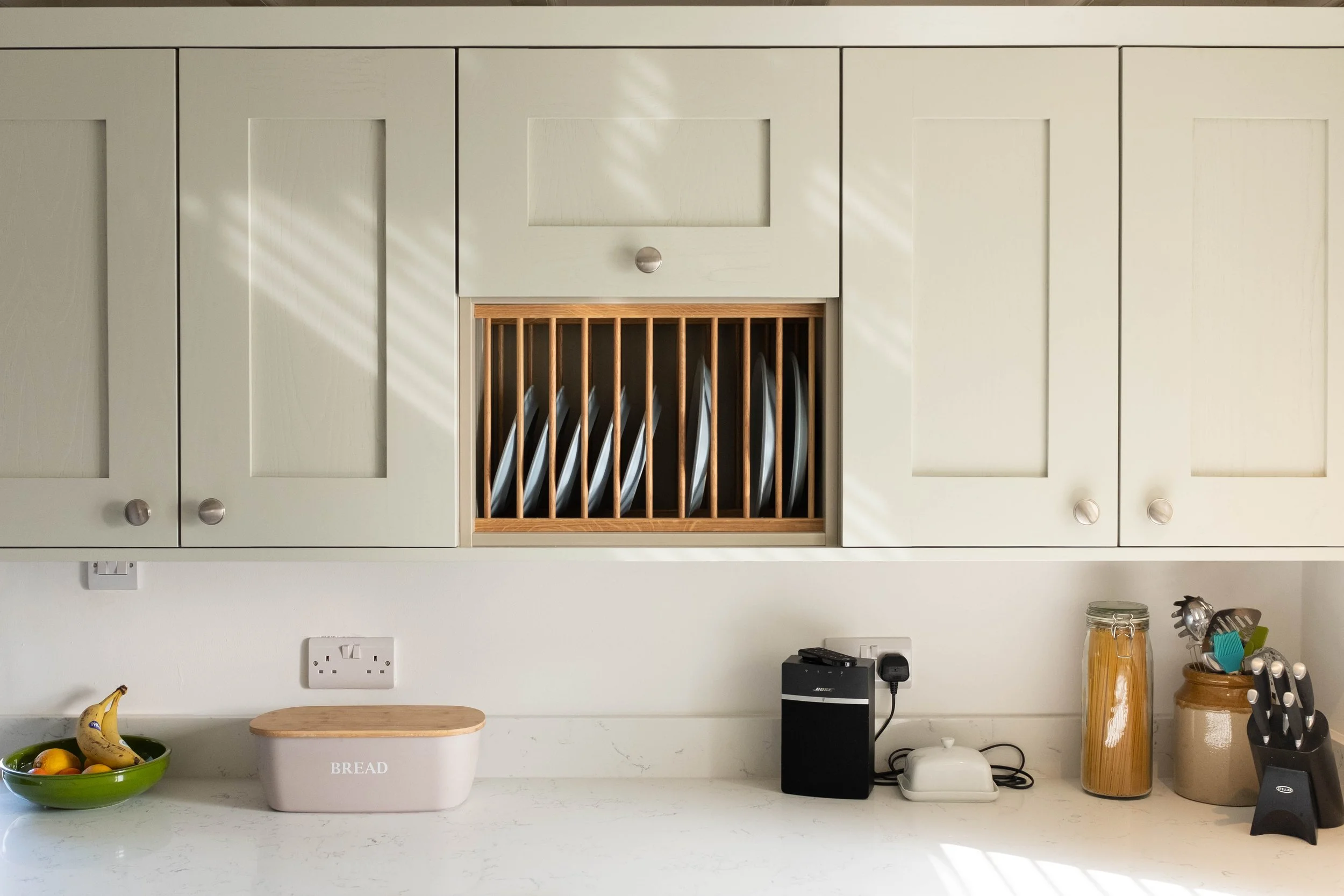 Kitchen wall with cream-colored cabinets and a wooden dish rack. Countertop has a white bread box, fruit bowl, small black speaker, toaster, jar with pasta, and utensil holder. Electrical outlets are visible.