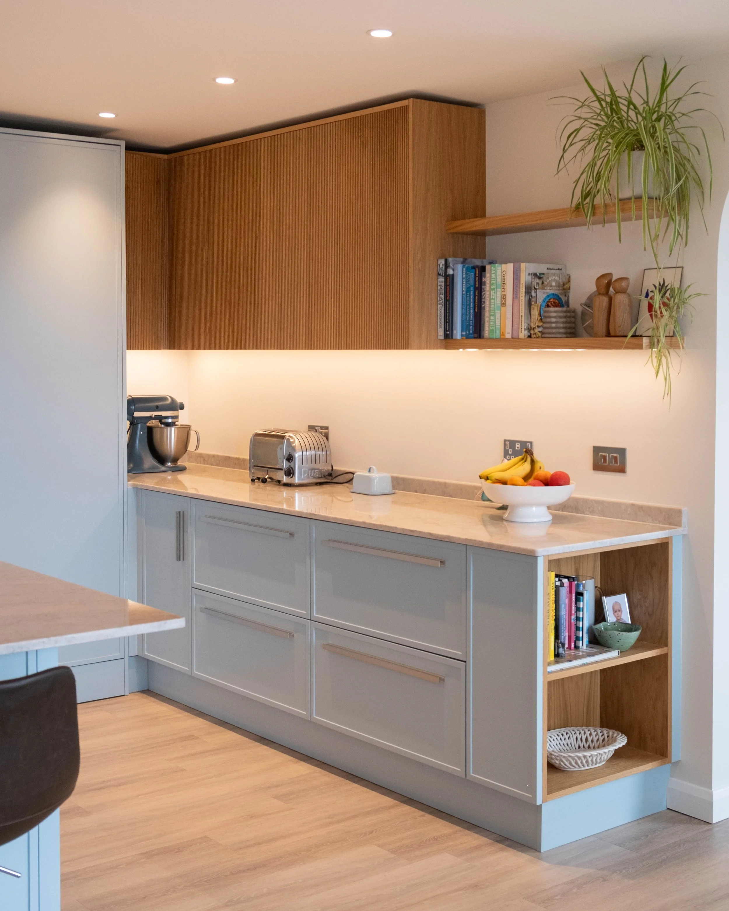 Kitchen with gray cabinets, wooden countertop, open shelving with books, plants, and decorative items, fruit bowl, and small appliances under ceiling lights.