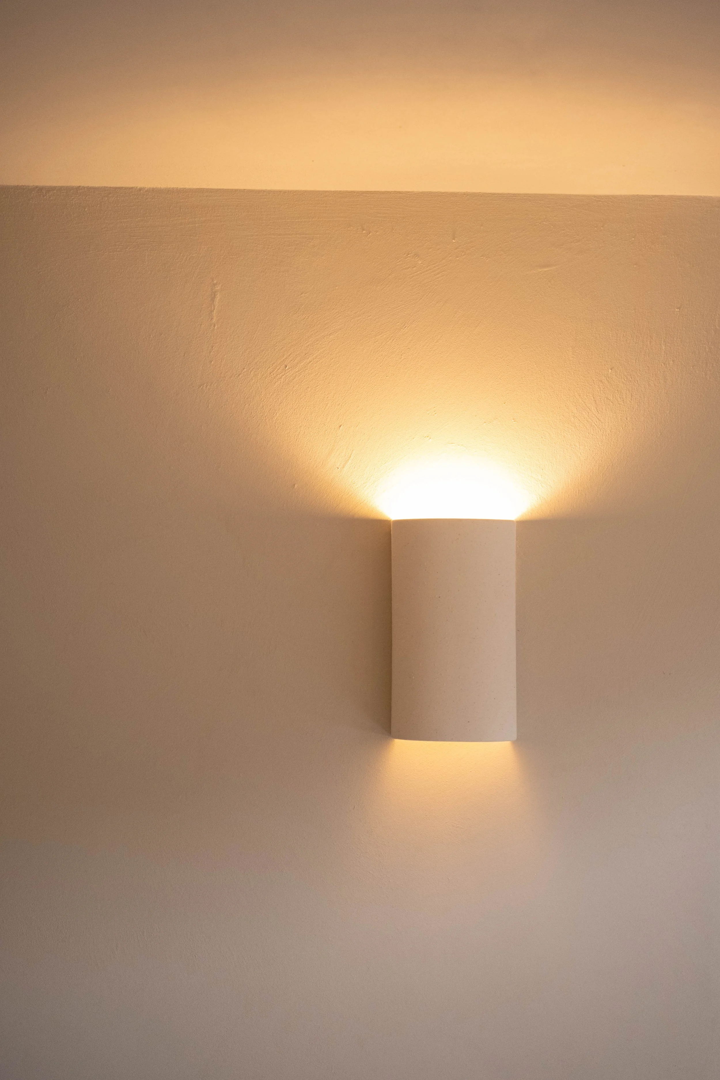 A modern wall sconce with a cylindrical white shade emits warm light onto a beige wall.