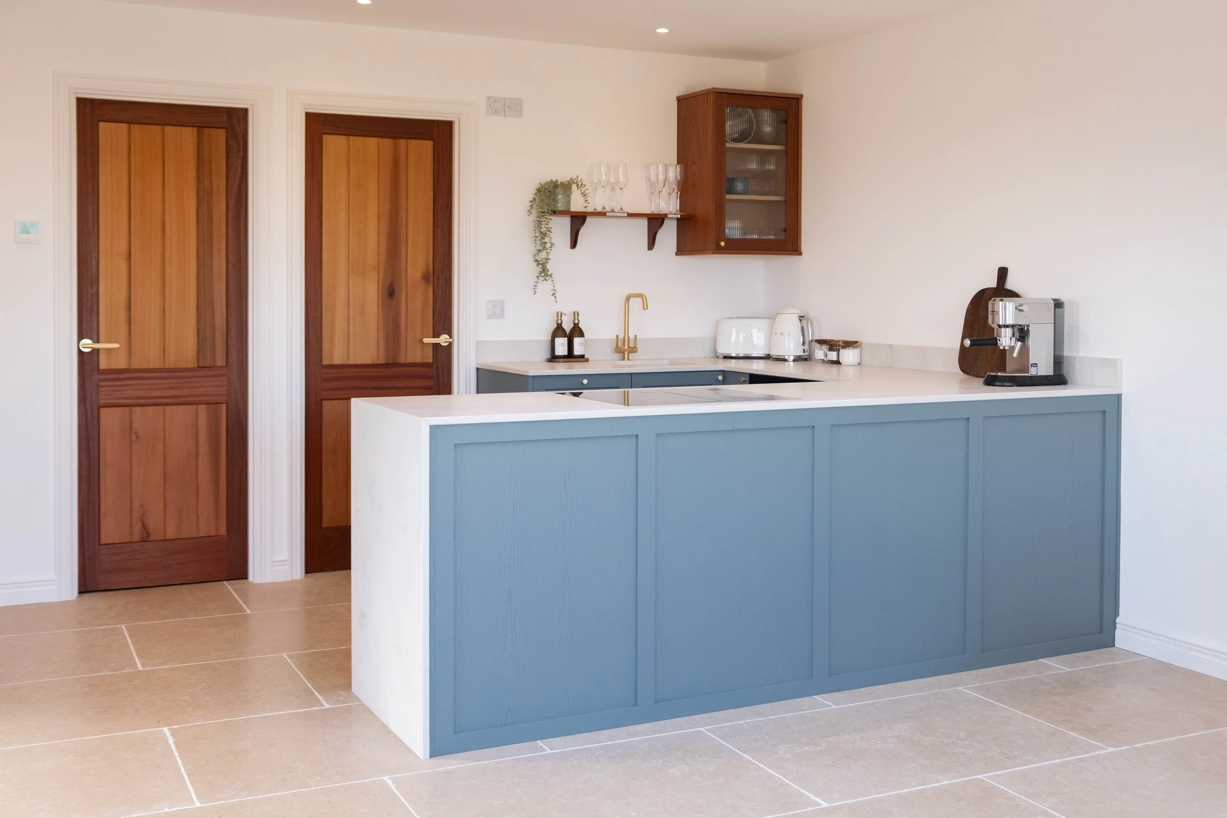 A kitchen with a blue island, wooden doors, and a white wall with cabinets and appliances.