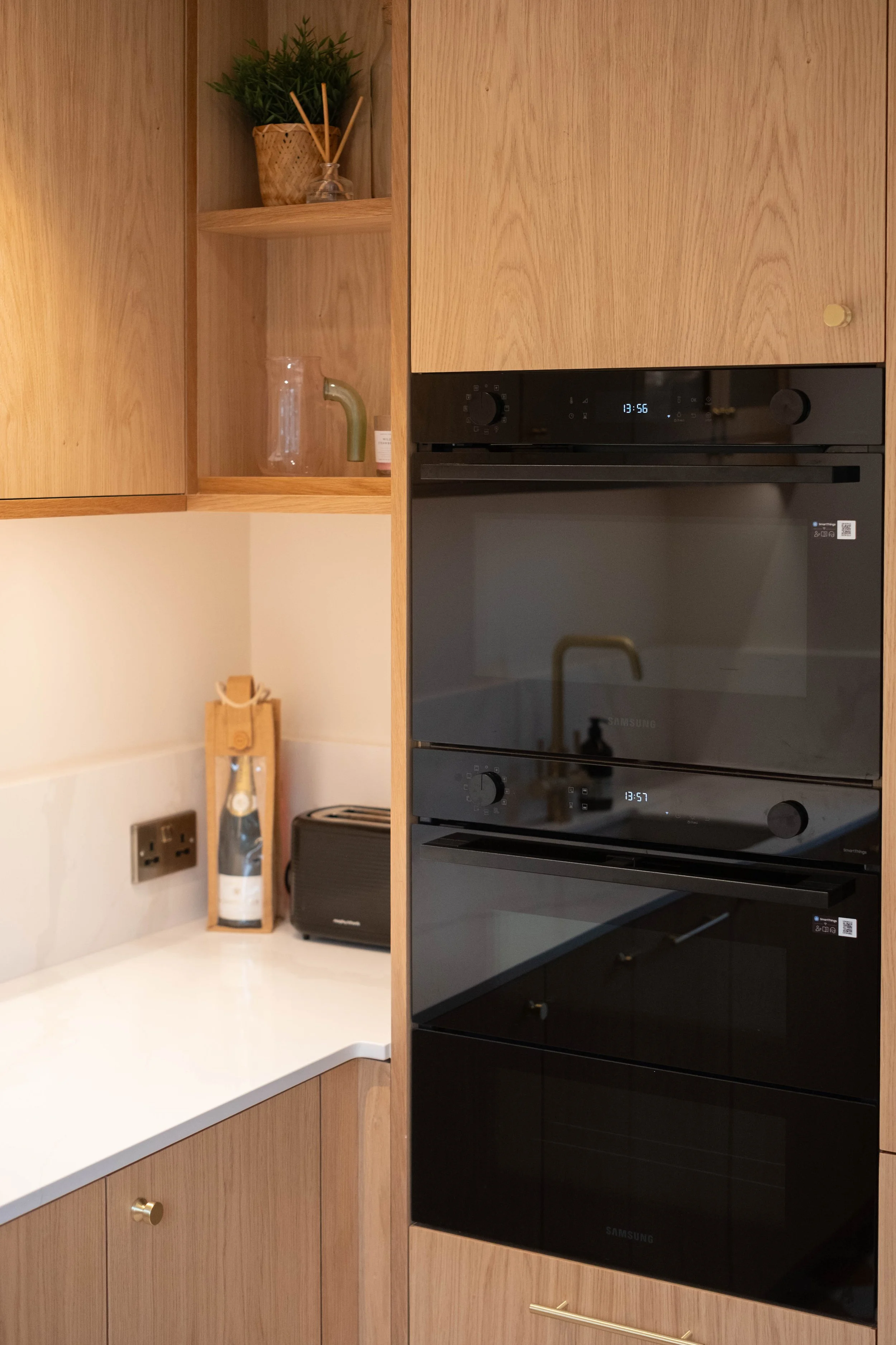 A modern kitchen with wooden cabinets, built-in black double oven, open shelf with a potted plant, a glass jug, and a small bottle, along with a white countertop, knife block, a water bottle, and a small black speaker.
