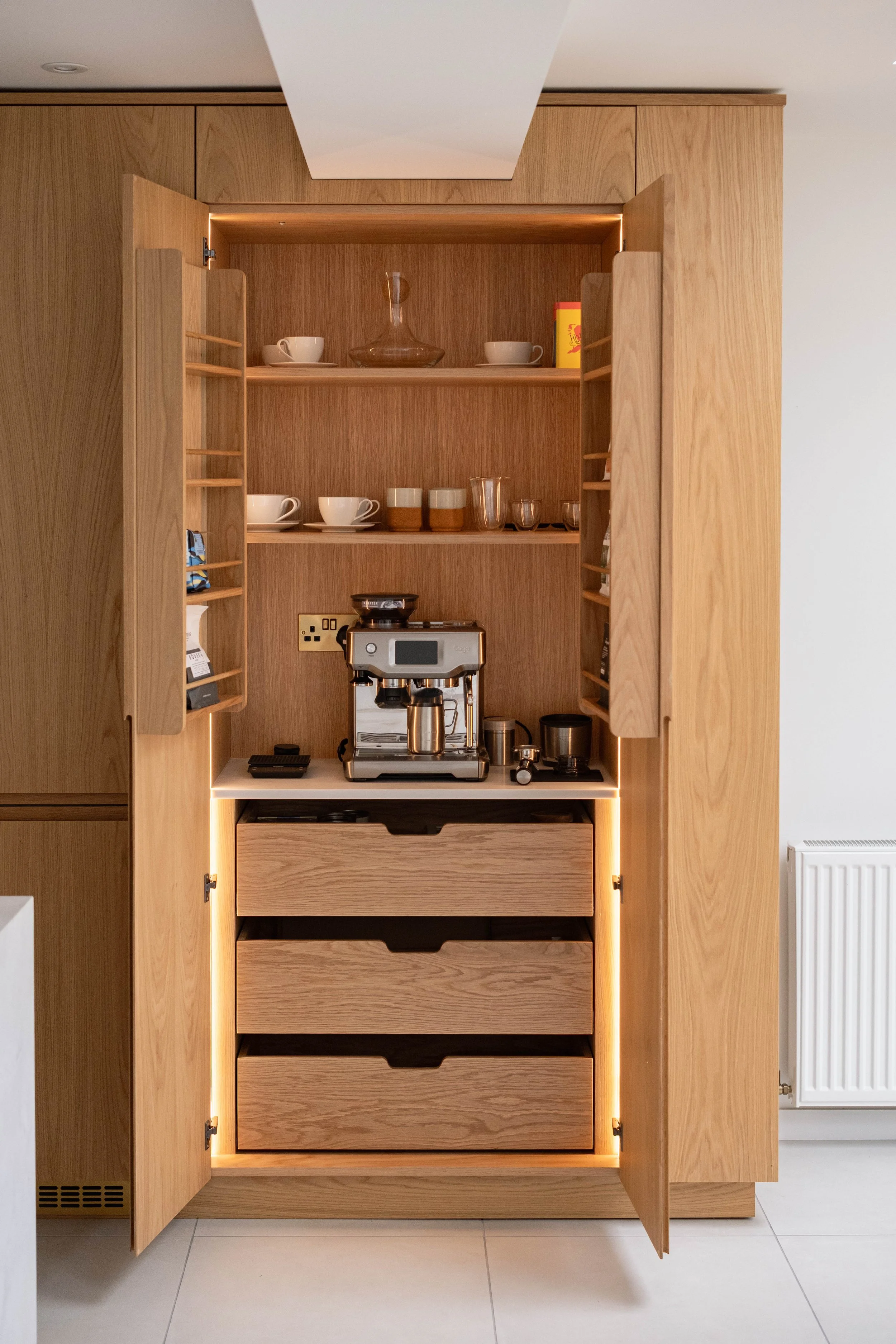 Open wooden kitchen cabinet with glassware, cups, and a coffee machine inside, against a wall with a radiator to the right.