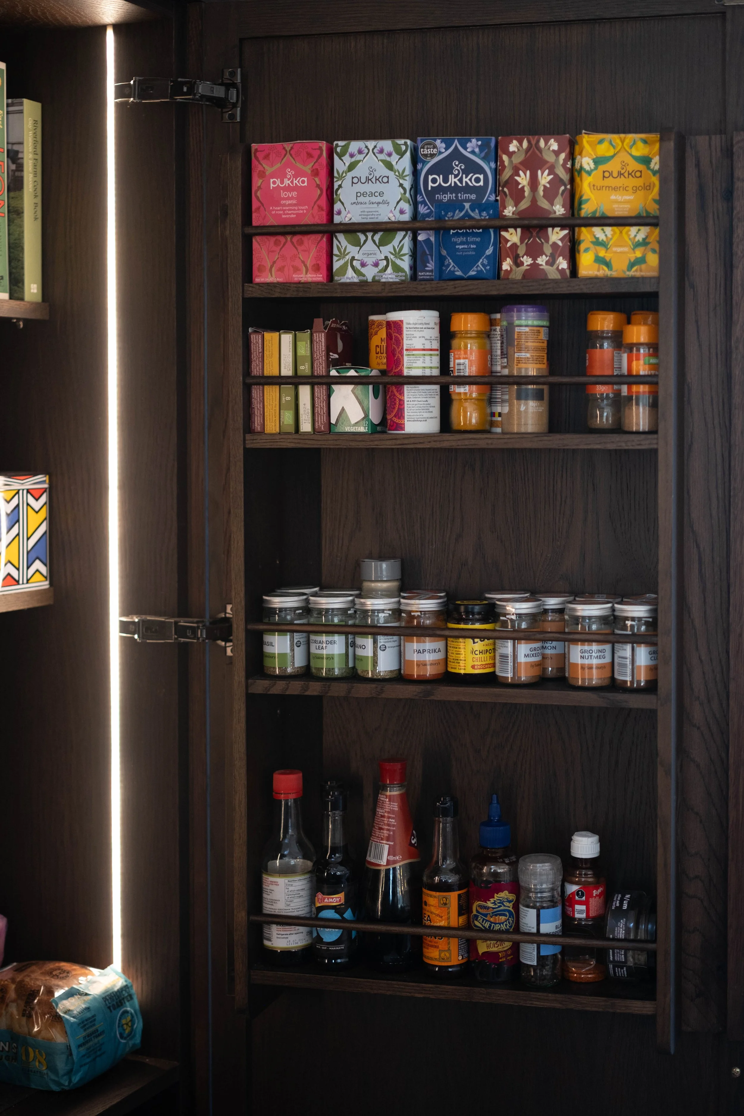 Dark wooden kitchen cabinet with three shelves filled with various spices, herbs, and condiments in bottles and jars. A bag of bread is visible on the bottom left corner.