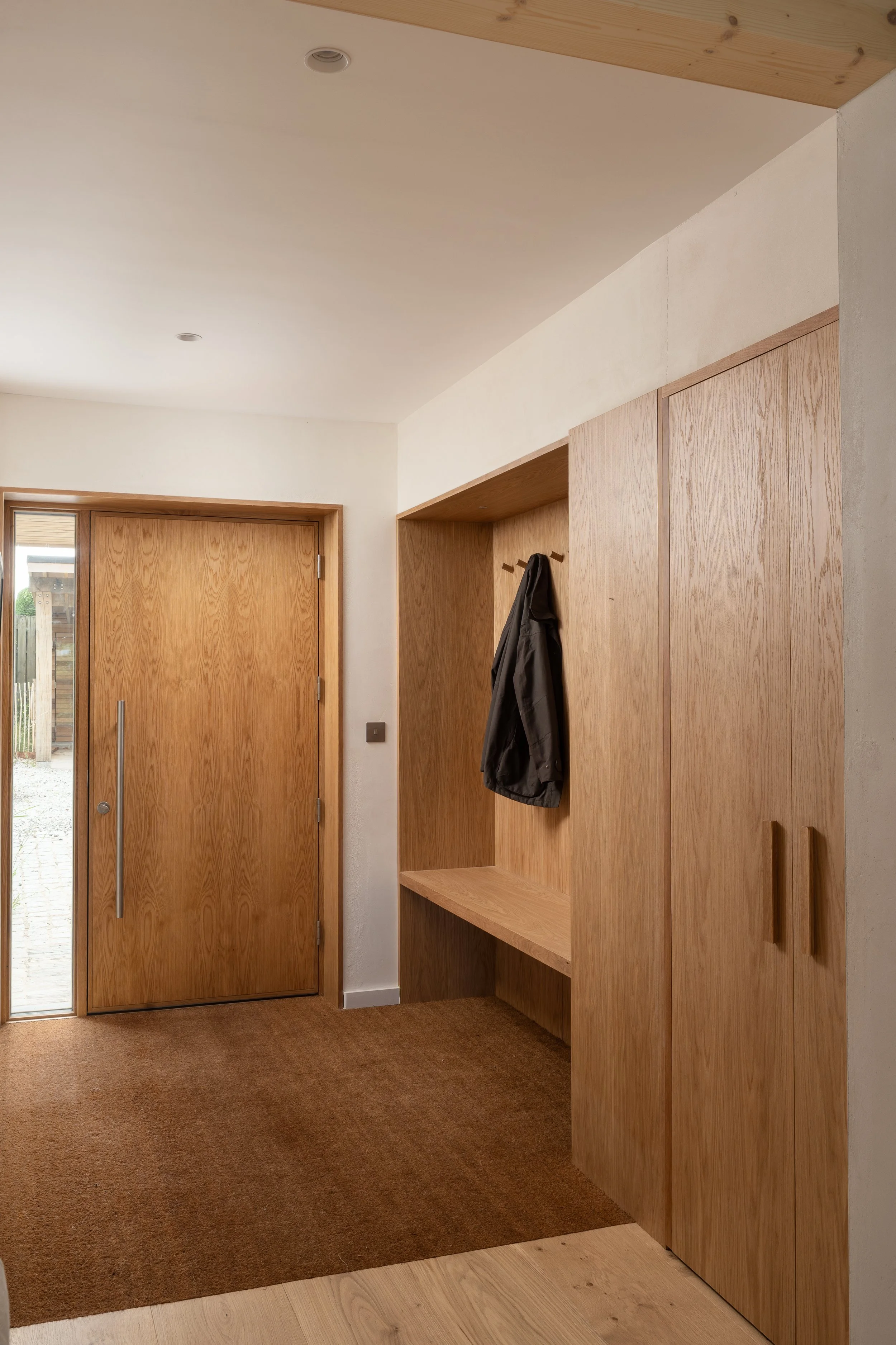 Interior of a modern house entryway with wooden doors, coat hooks, and a built-in wooden storage cabinet, with ceiling lights and a brown carpet carpeted floor.