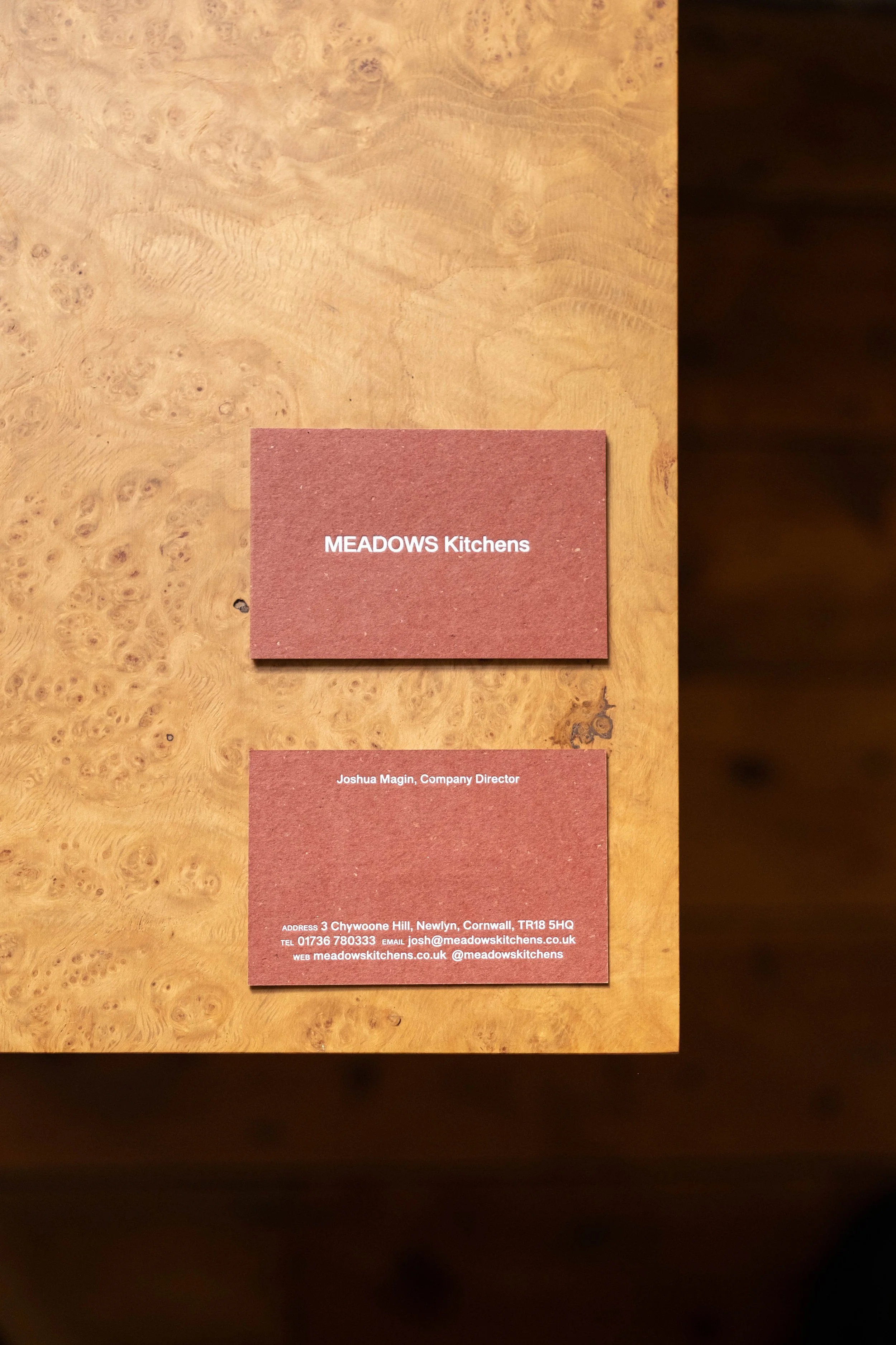 Two business cards on a wooden desk. The top card reads 'MEADOWS Kitchens.' The bottom card contains contact information for Joshua Magin, Company Director, with address, phone number, email, and website details.