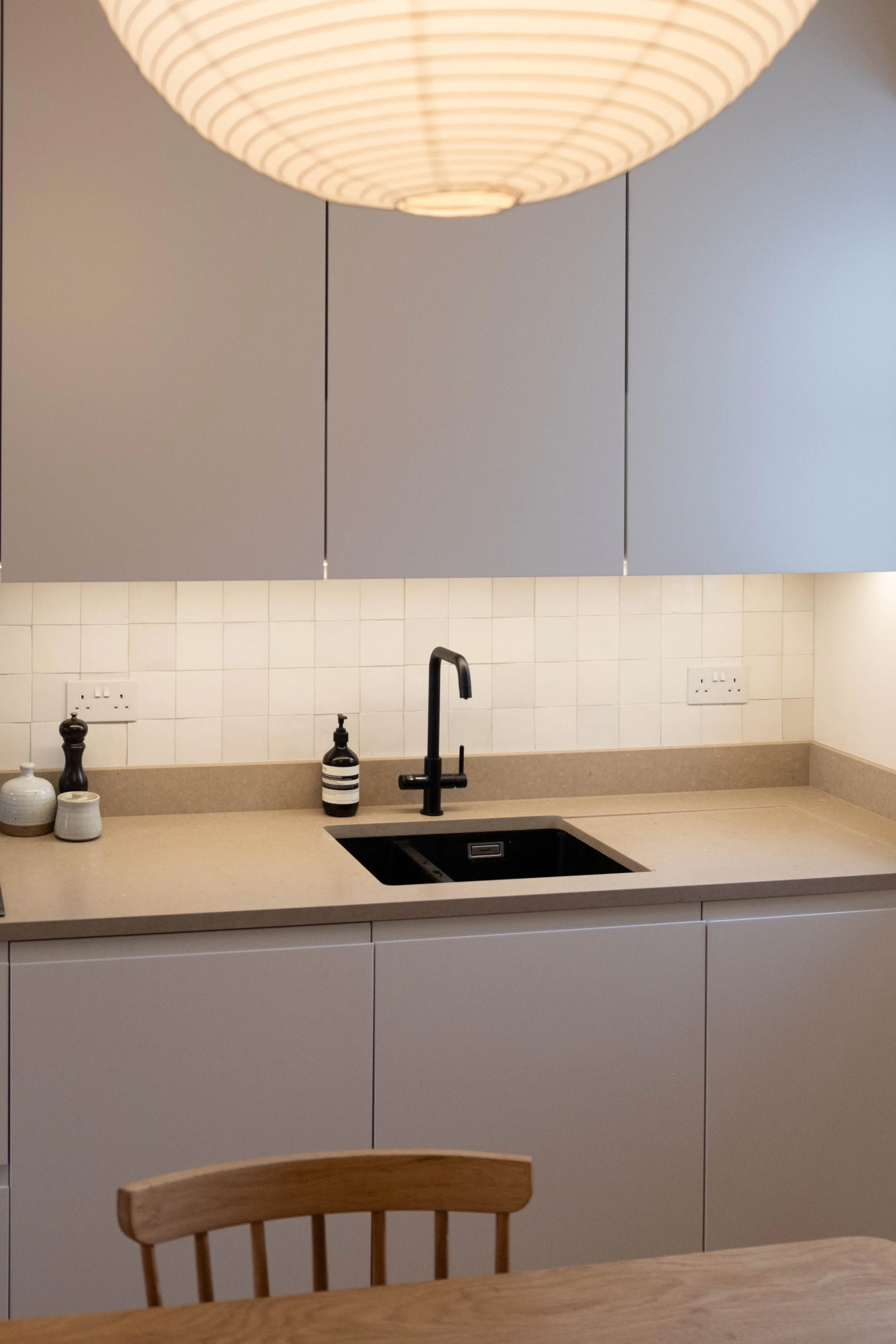 A modern kitchen with a beige countertop, black sink with a matte black faucet, white tiled backsplash, and upper cabinets painted in gray. There is a wooden dining chair and decorative ceramic jars on the countertop, with a large, round, white penda