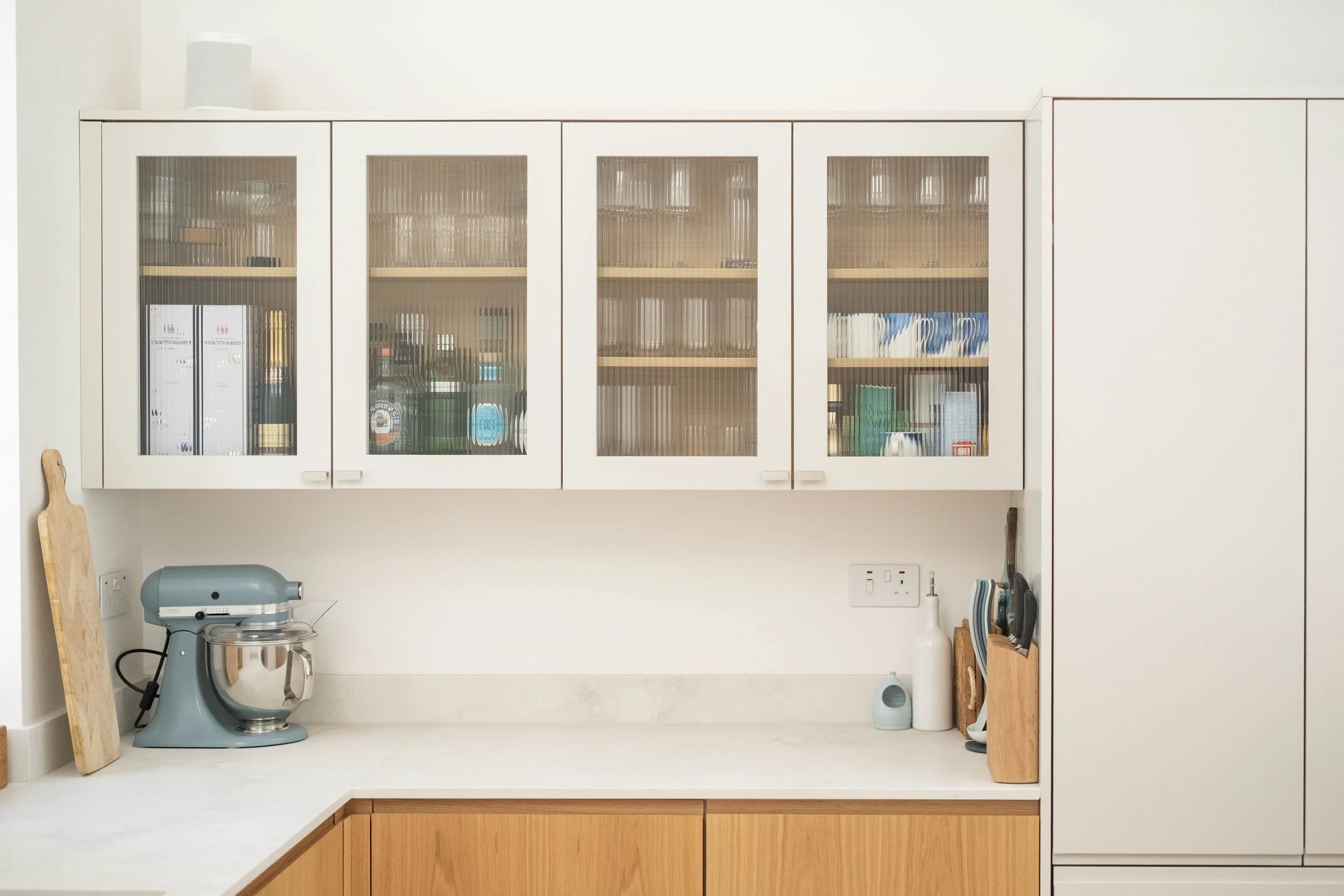 A white kitchen wall with a cabinet having glass doors, containing binders, books, and jars. Below, there is a countertop with a grey stand mixer, a wooden cutting board, and various kitchen utensils. An electrical outlet and a soap dispenser are als