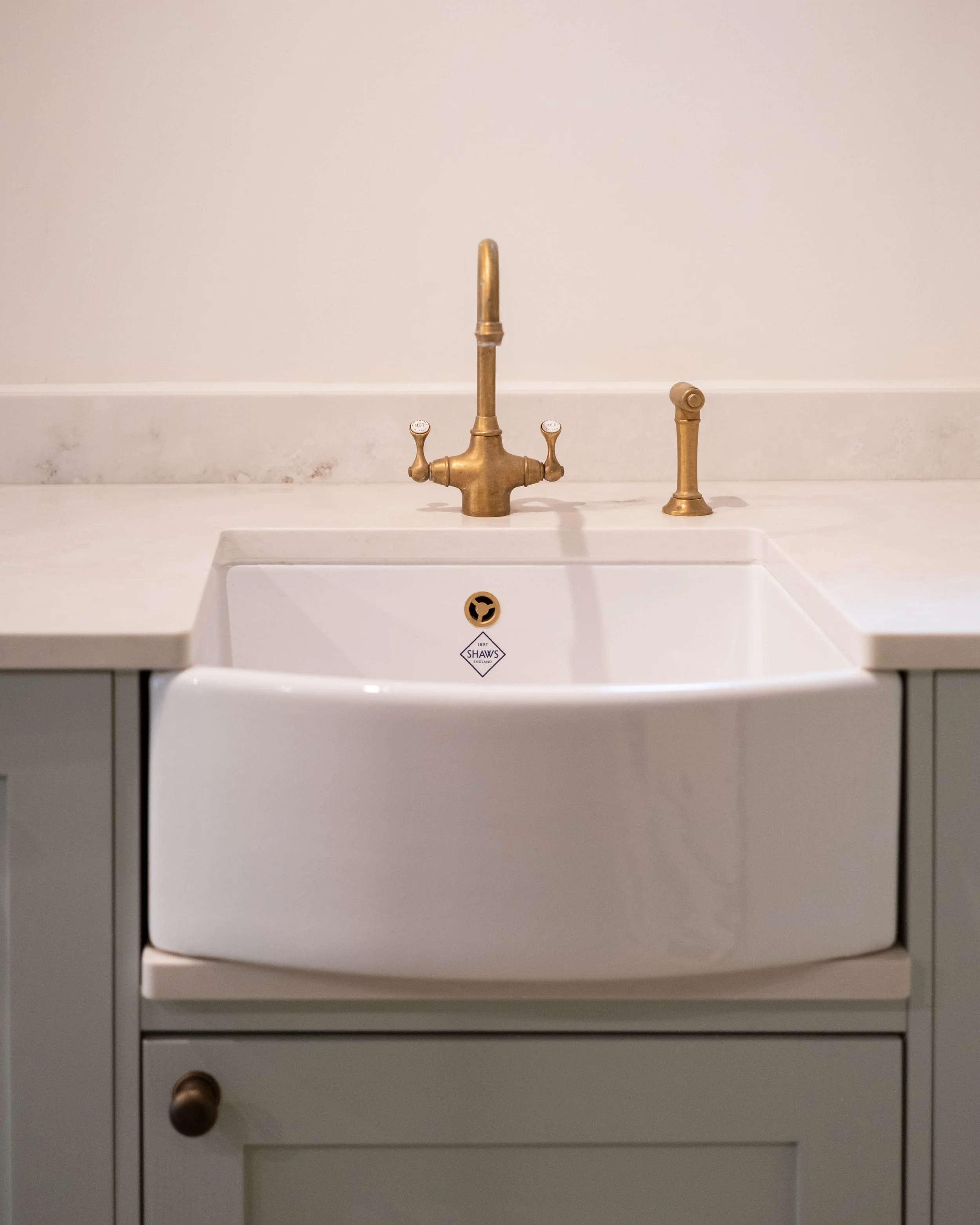 A white kitchen sink with a gold-colored faucet and a separate gold-colored knob. The background is a plain, light-colored wall.