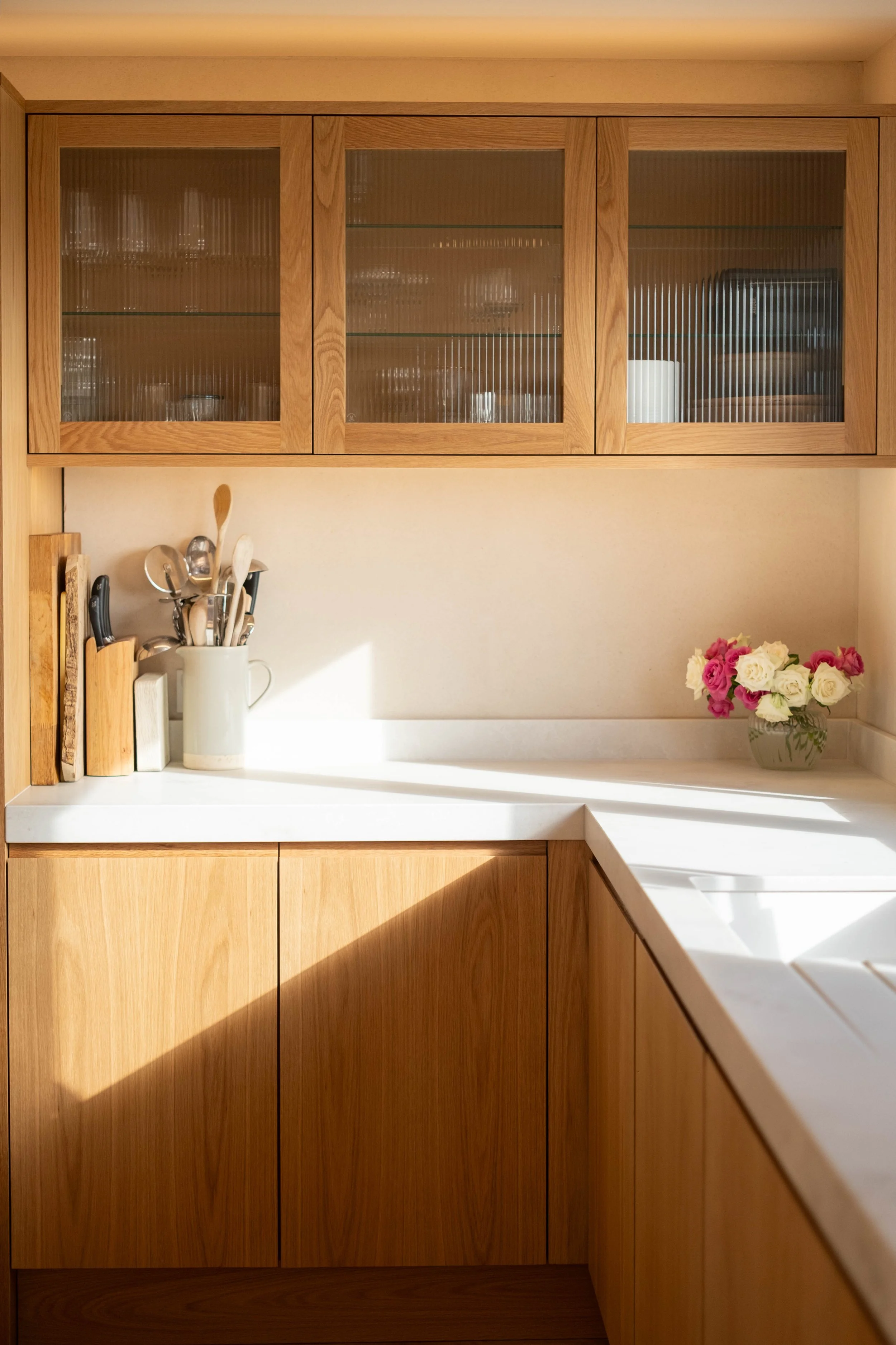 Wooden kitchen cabinets with glass doors, a white mug holding kitchen utensils, and a vase of pink and white flowers on a white countertop.