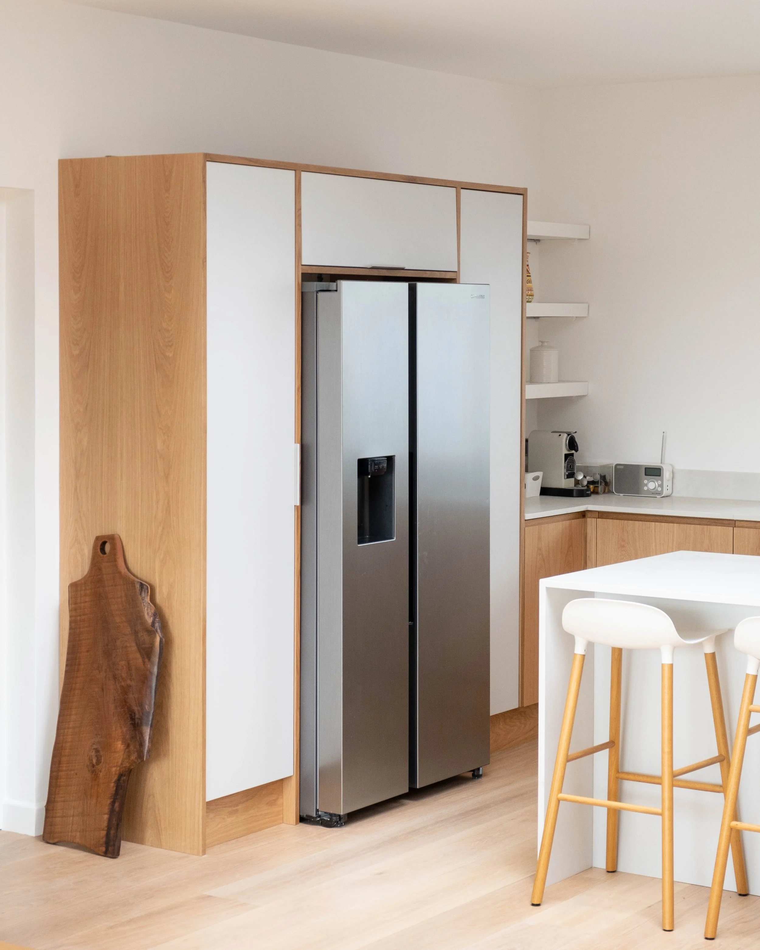 A modern kitchen with light wood flooring, white walls, and minimalistic design. Features include a wooden board leaning against the wall, a built-in kitchen with a stainless steel refrigerator, and a white kitchen island with bar stools. Two white s