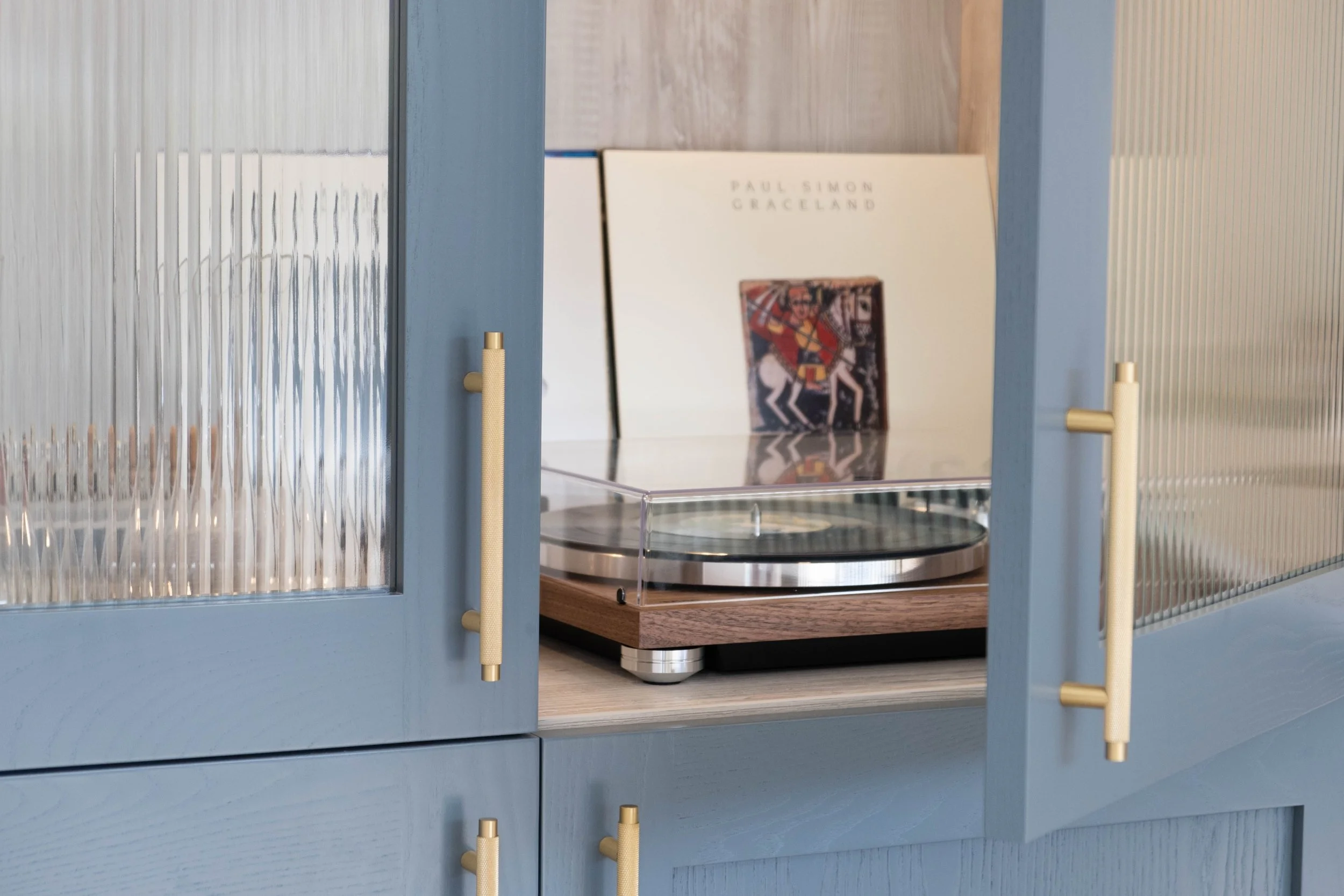 Close-up of a turntable with a vinyl record, partially visible behind a glass display case, with a white wall and artwork in the background.