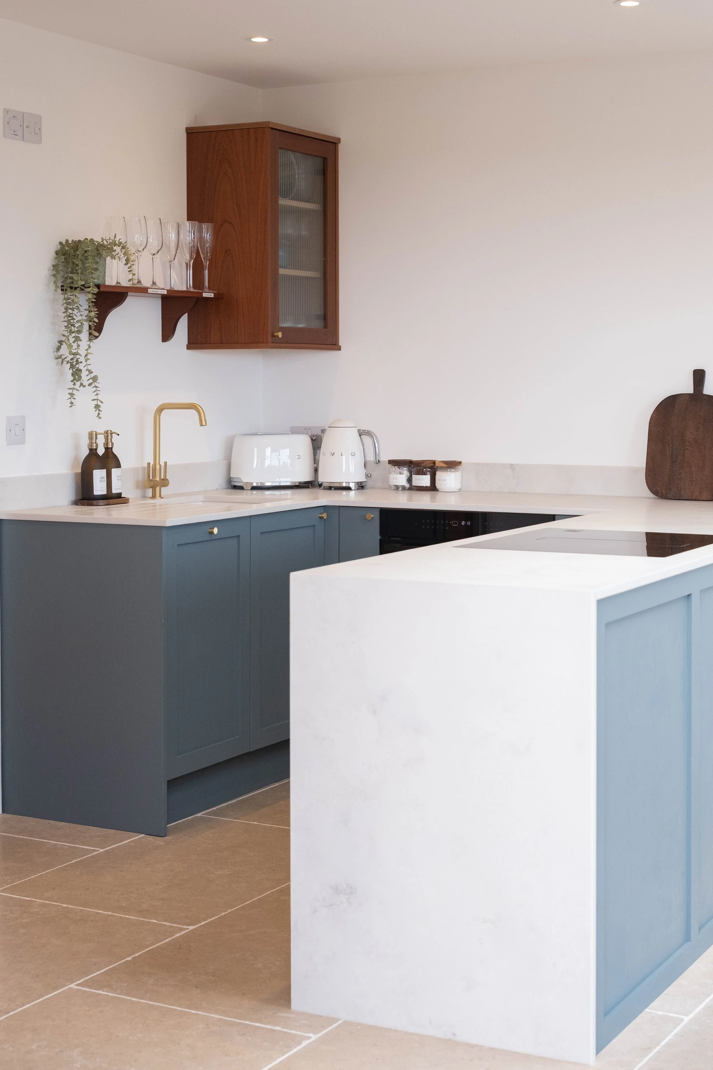 A modern kitchen with blue lower cabinets, a white countertop, a gold kitchen faucet, and small appliances including a toaster and kettle. A wooden wall-mounted cabinet and a shelf with glassware and a plant are also visible.