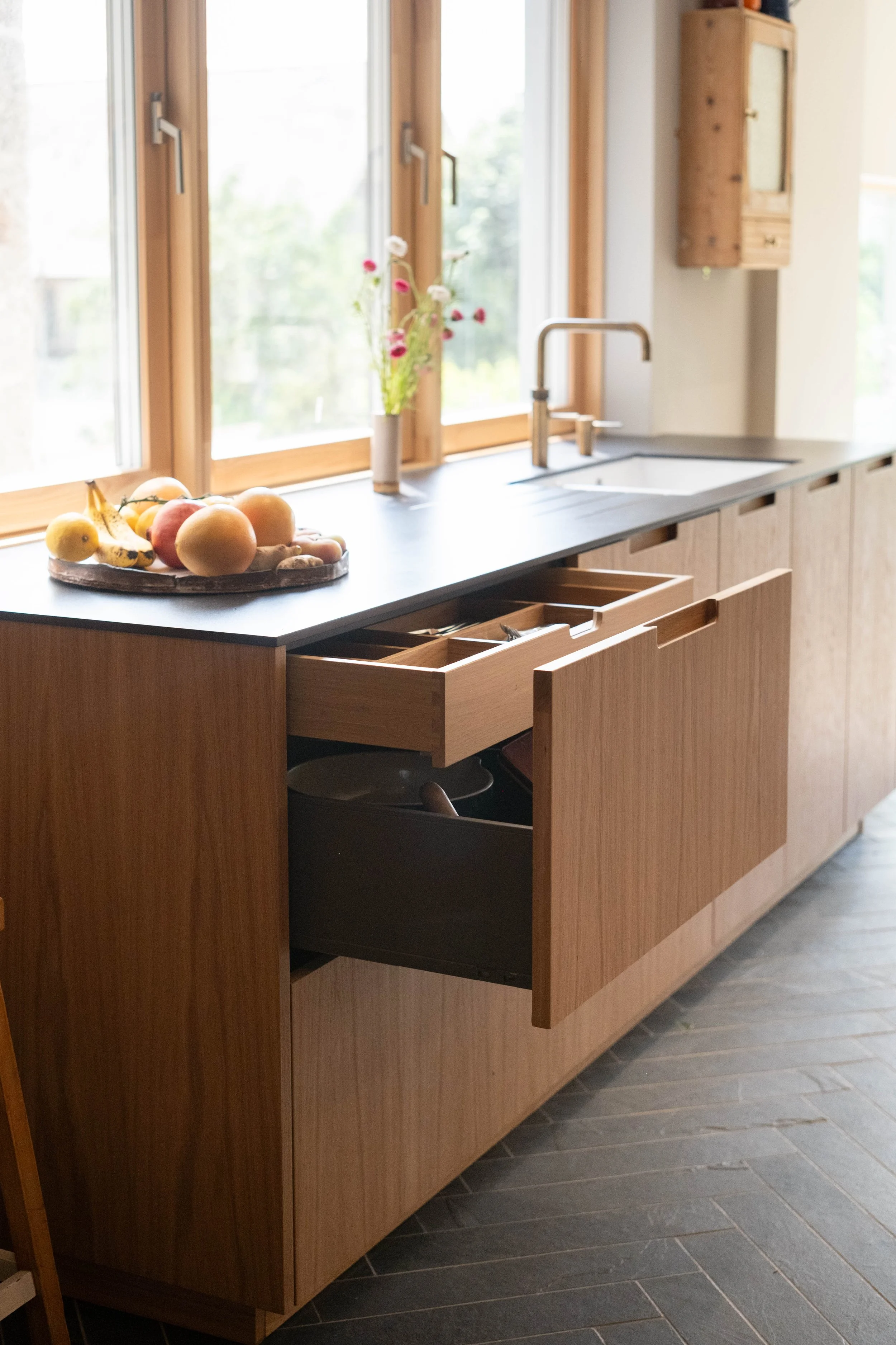 A kitchen with a wooden cabinet and a black countertop. On the countertop, there's a wooden tray with bananas, apples, and peaches. Behind the tray, a window with wooden frames and a view of trees outside. Next to the window, a vase with pink and whi