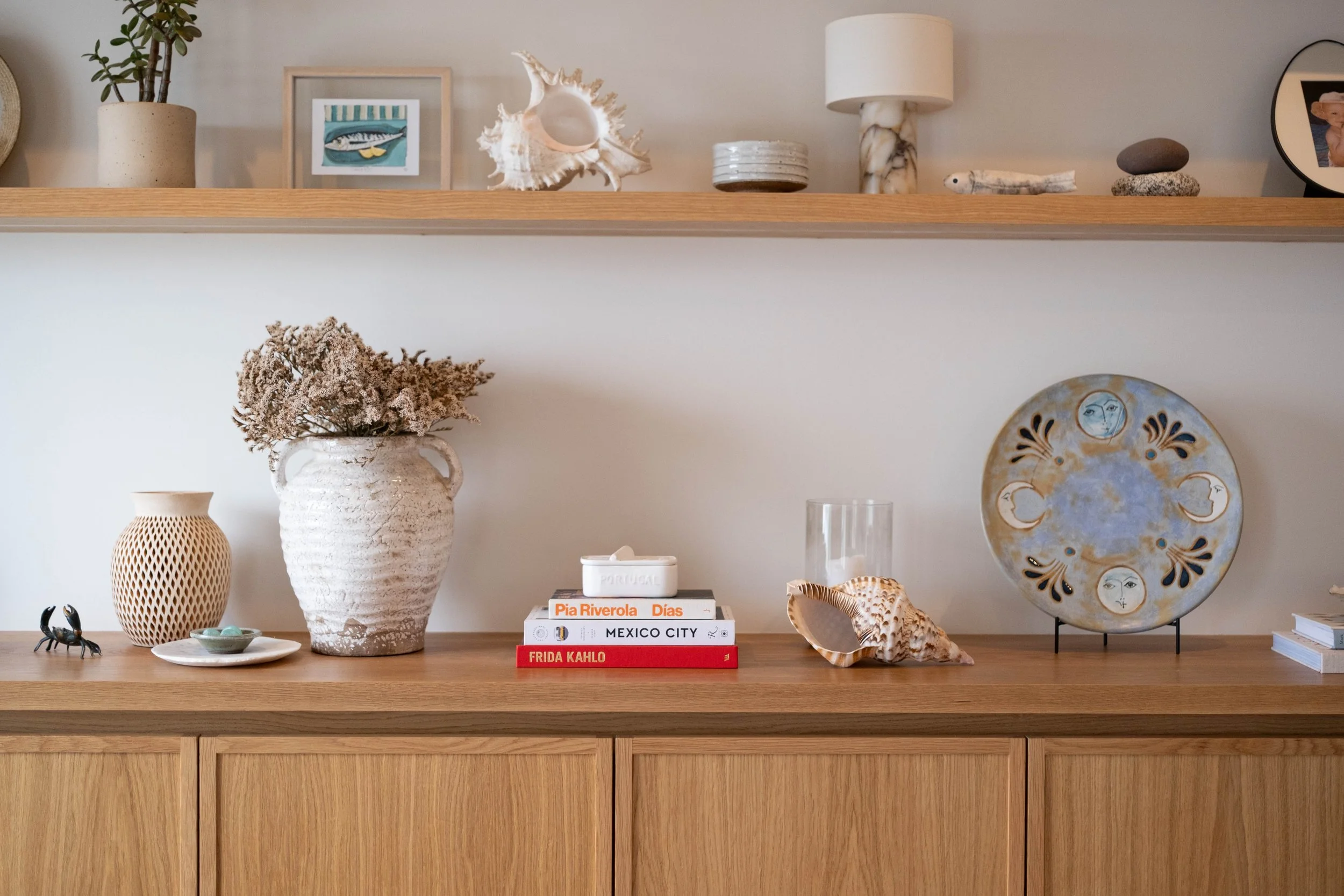 A wooden sideboard with decorative items including a large textured white vase with dried flowers, a beige ceramic vase with a lattice pattern, a small black crab figurine, a white dish with turquoise stones, a stack of books with titles about cities