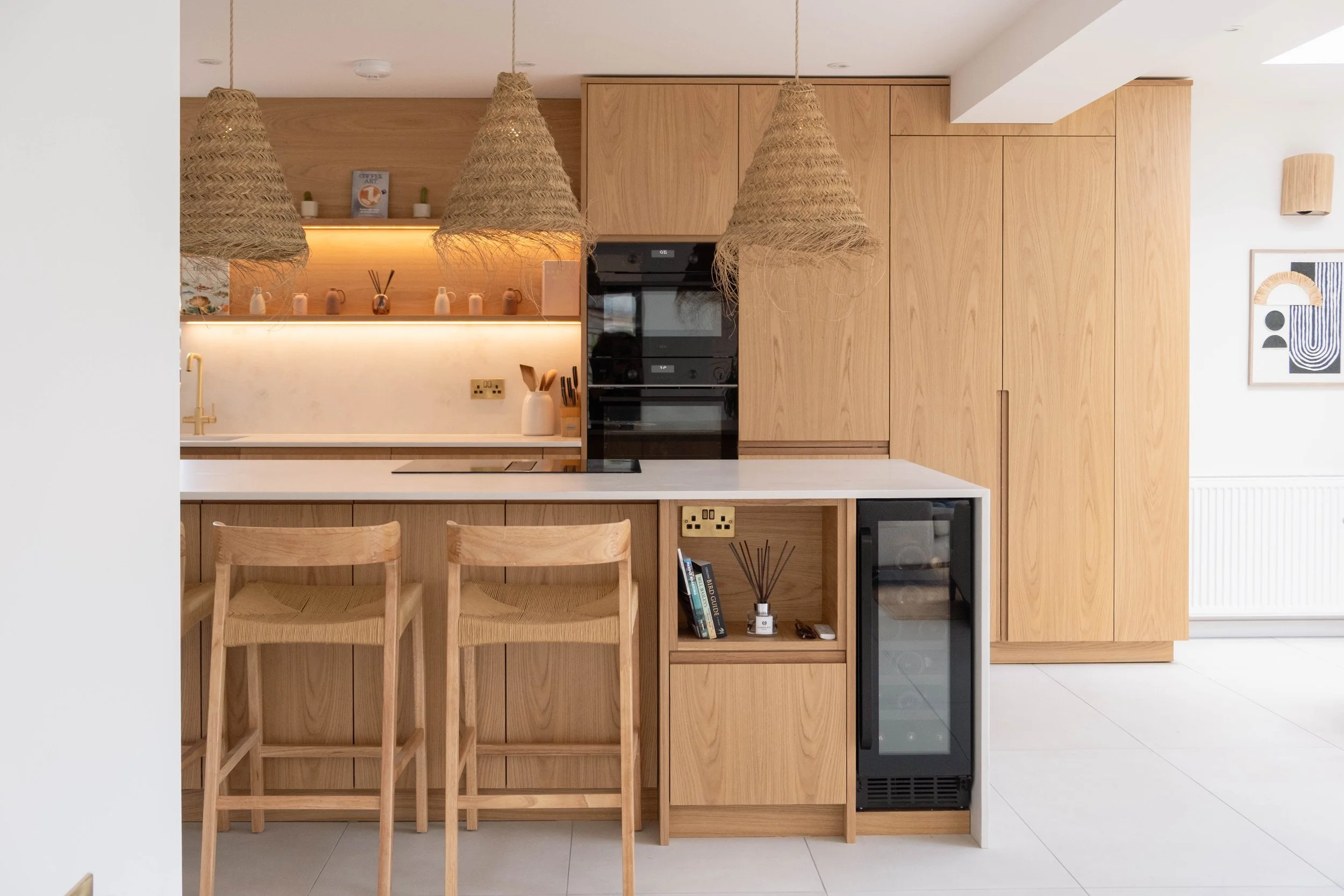 A modern kitchen with wooden cabinets, a white countertop, three wooden bar stools, pendant lights made of woven material, and decorative items on a shelf in the background.