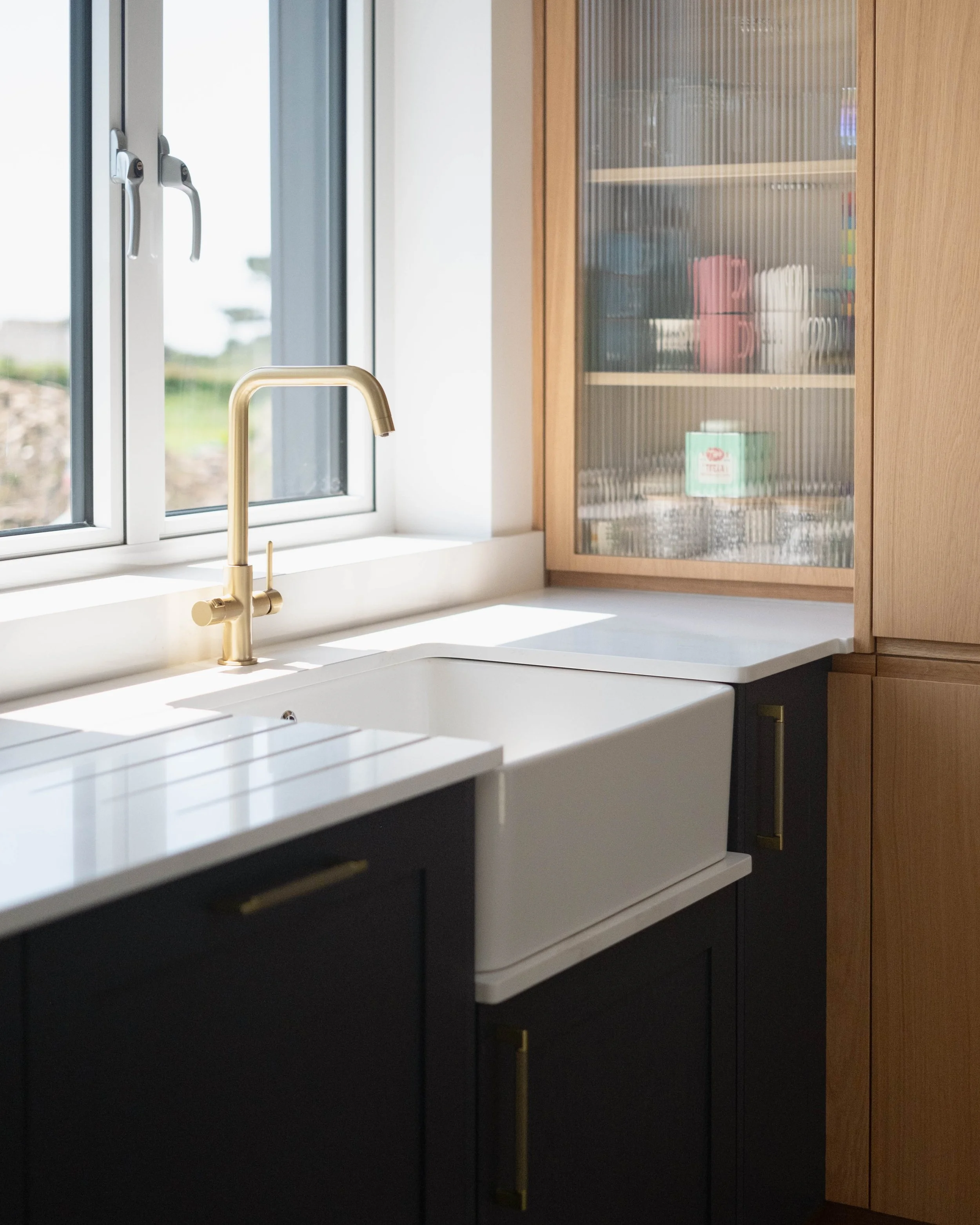 View of a modern kitchen sink with a gold-colored faucet next to a window, a cabinet with a glass door showing various household items inside, and part of a drawer below the sink.