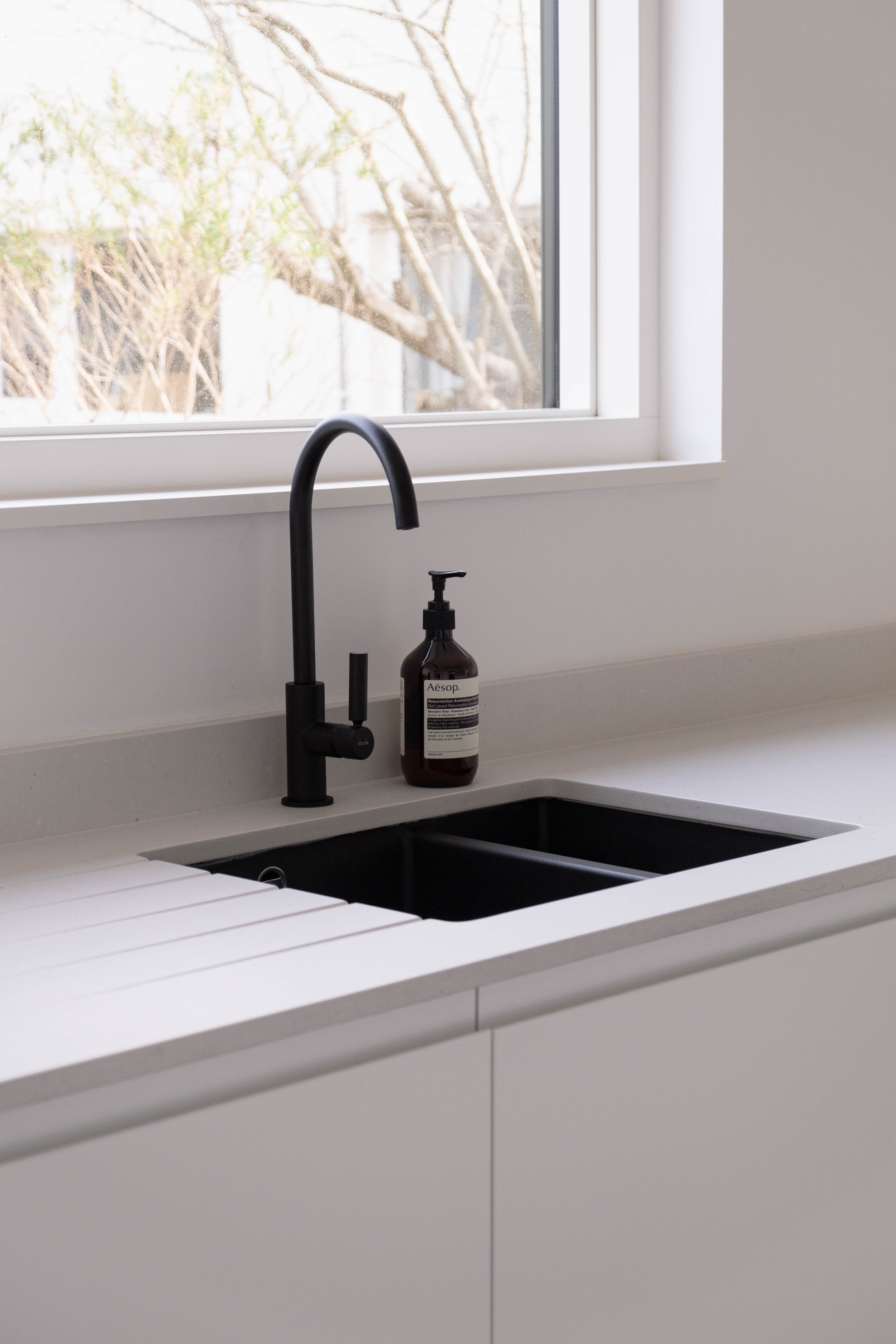 A modern kitchen with a white countertop, black sink, black faucet, and a brown soap dispenser labeled "Aesop." There is a window above the sink showing a sunny outdoor view with trees.