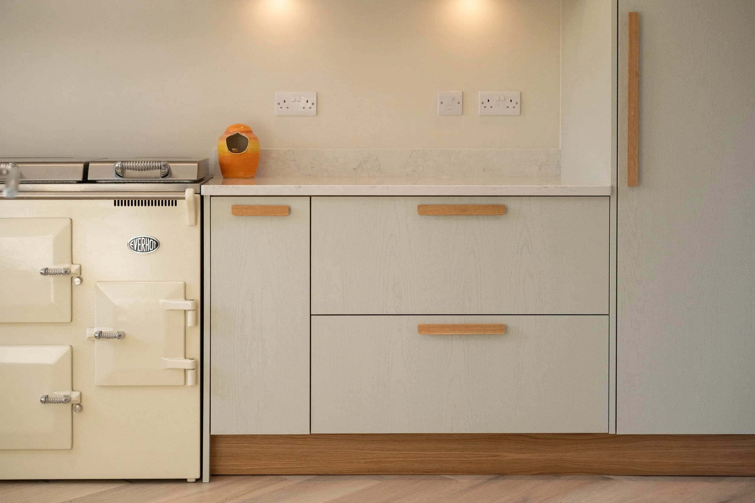A kitchen countertop with electrical outlets on the wall, a decorative orange vase, and light-colored wooden cabinets and drawers.