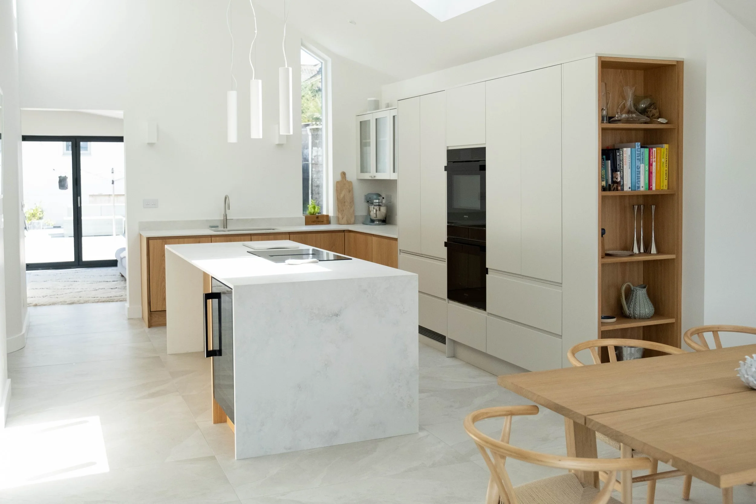 Modern kitchen with white cabinets, a white island, a wooden dining table with matching chairs, and open shelving with books and decorative items, illuminated by natural light from windows and a skylight.