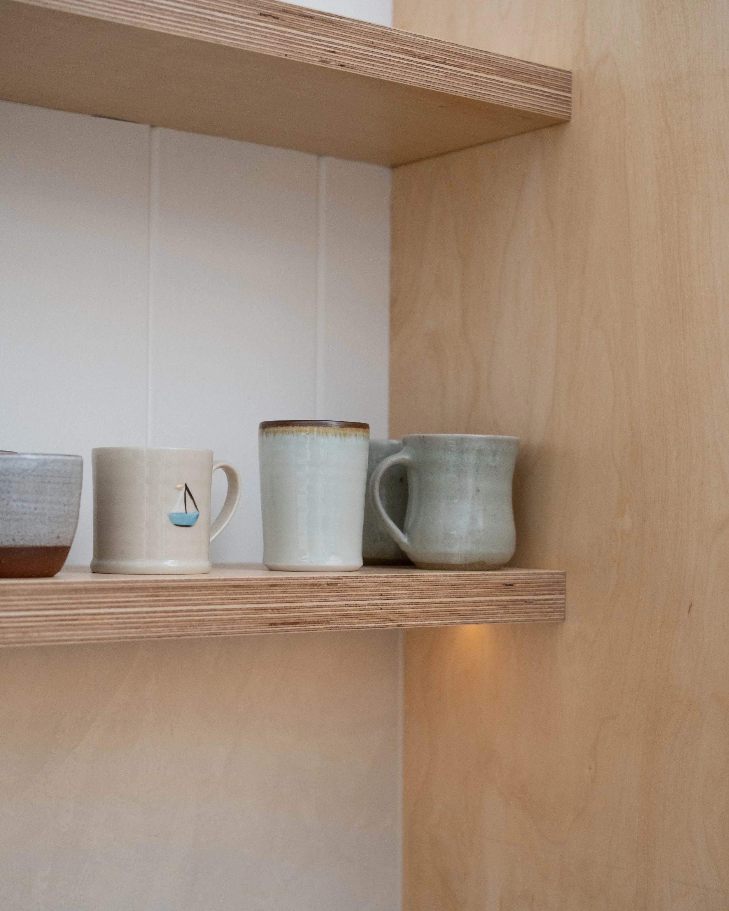 Close-up of four ceramic mugs on a wooden shelf, with various textures and colors, against a plain wall.