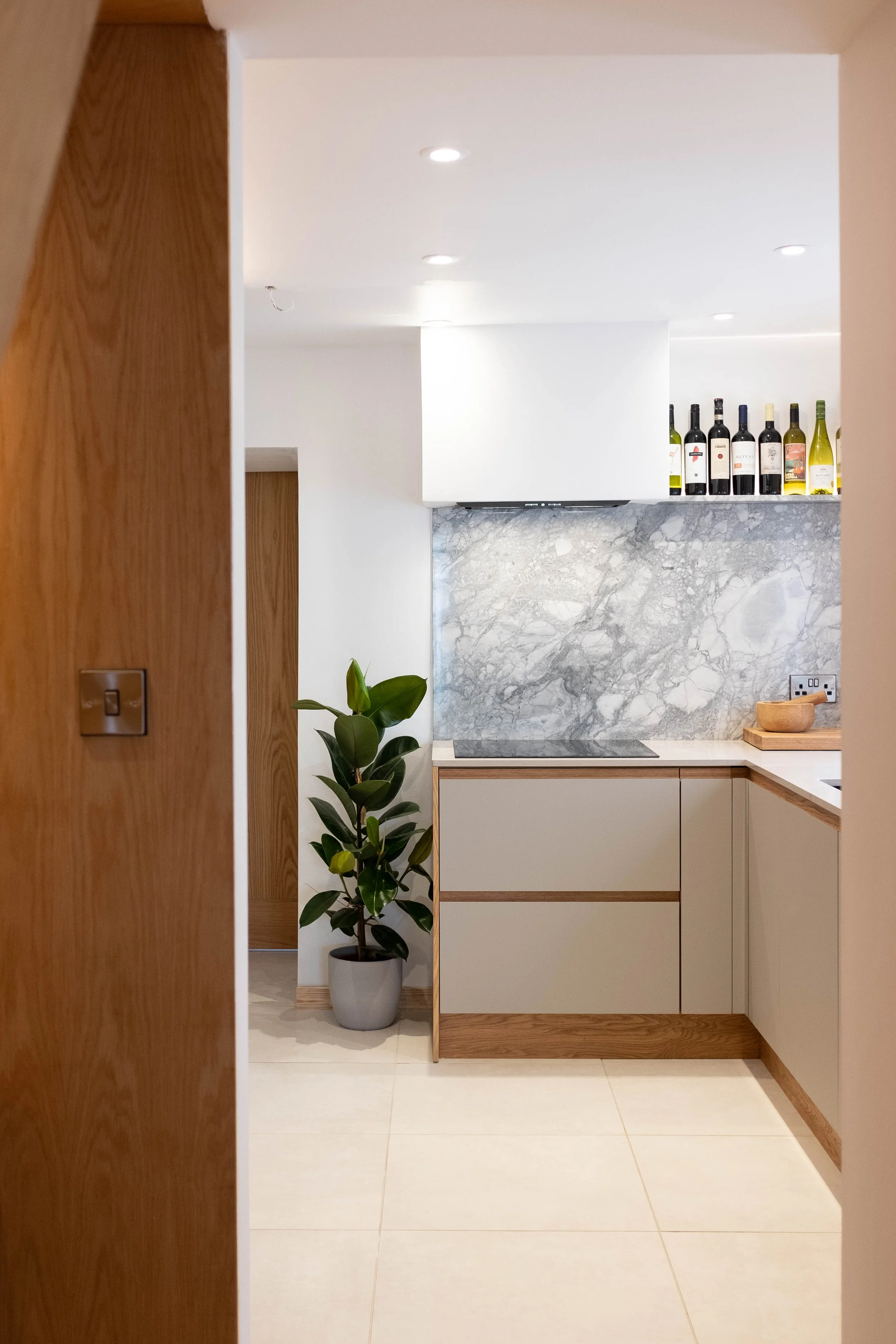 A modern kitchen with white cabinets, a grey marble backsplash, and a counter with a wooden bowl. A potted plant is situated on the floor near the cabinets. On the upper shelf, there are several bottles of wine against a white wall. The flooring is l