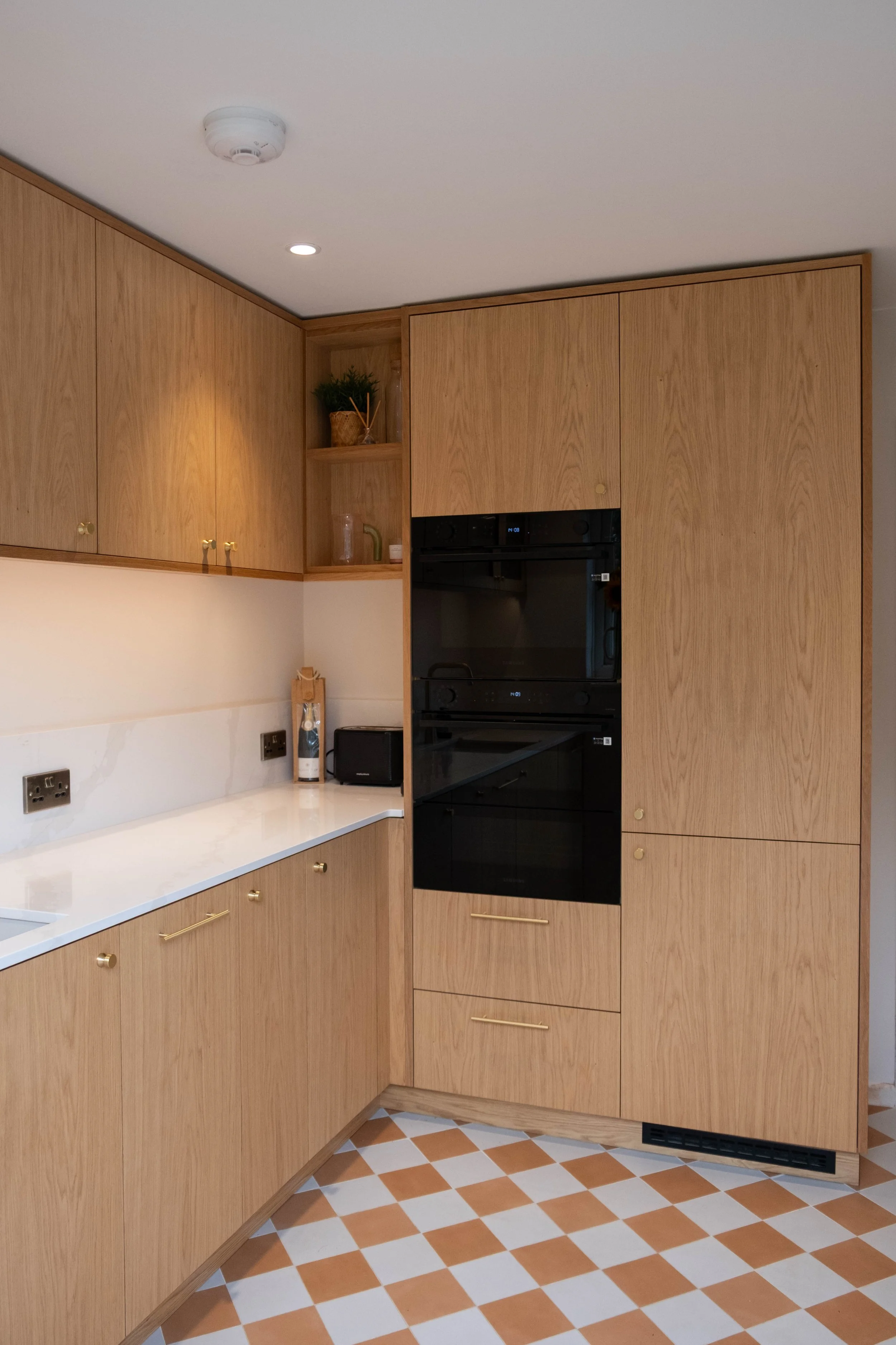 A kitchen corner with wooden cabinets, a white countertop, and a built-in black oven. The floor has a checkered orange and white tile pattern. There are small decorative items and a plant in one open shelf.