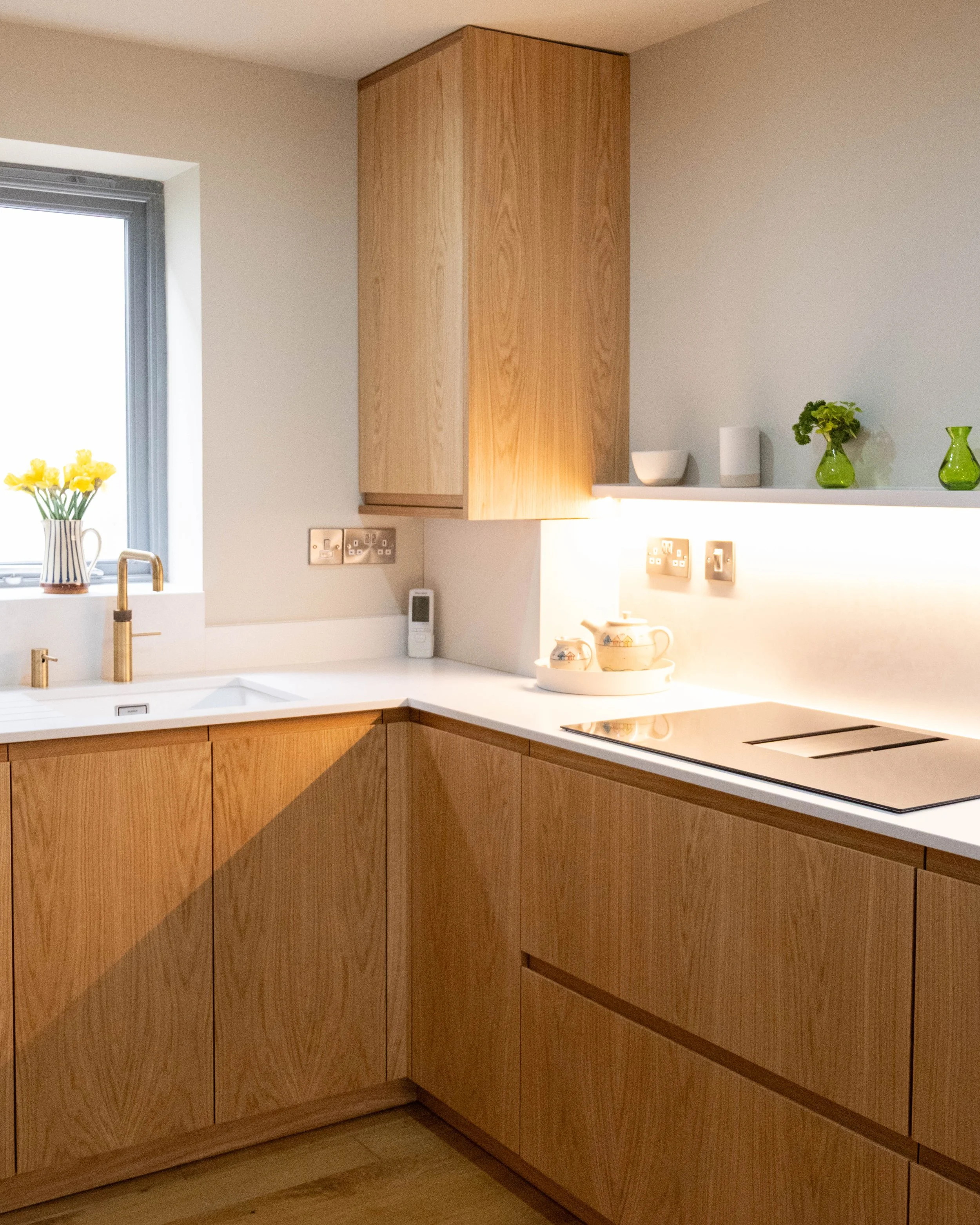 Wooden kitchen cabinets with a white countertop, a window with a vase of yellow flowers, and decorative items including green vases and bowls.