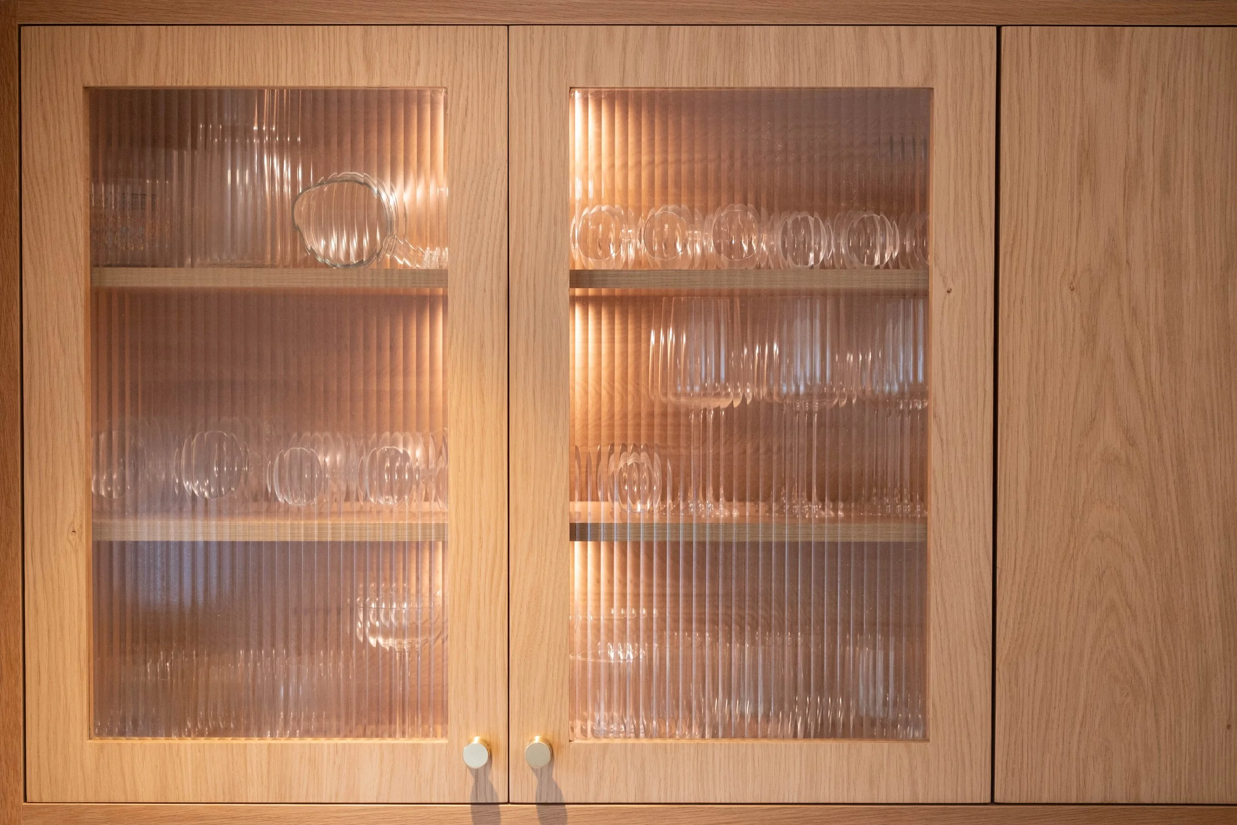 Wooden cabinet with glass-paneled doors and round handles, containing glassware such as drinking glasses and bowls inside.