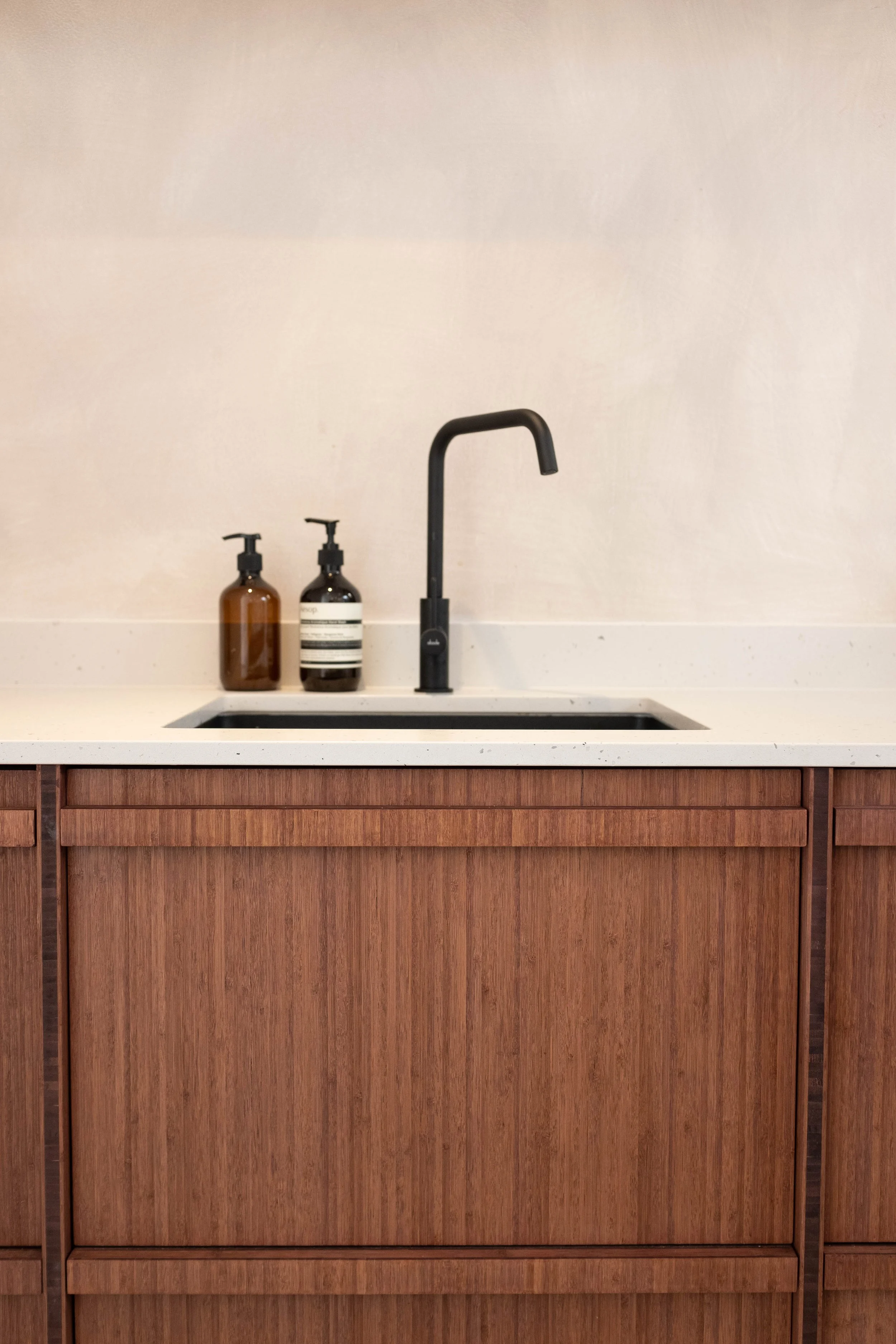 Kitchen sink with black faucet and two bottles of soap or lotion on a white countertop against a beige wall with wood cabinetry.