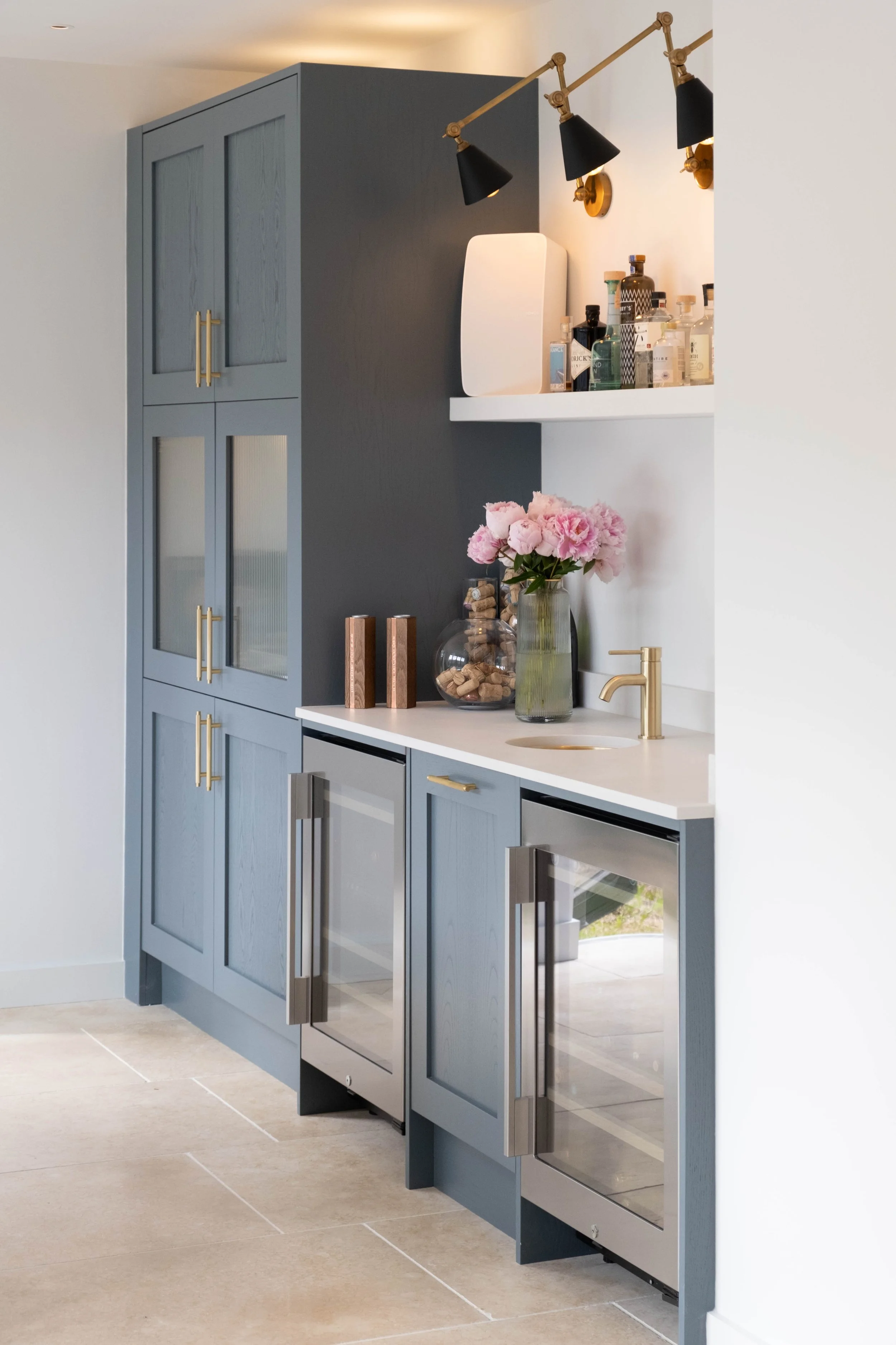 Blue kitchen cabinet with gold handles, a white countertop with a sink and gold faucet, a glass vase with pink flowers, a transparent jar filled with wine corks, and a white shelf with various bottles. Black and gold wall-mounted lamps above the shel