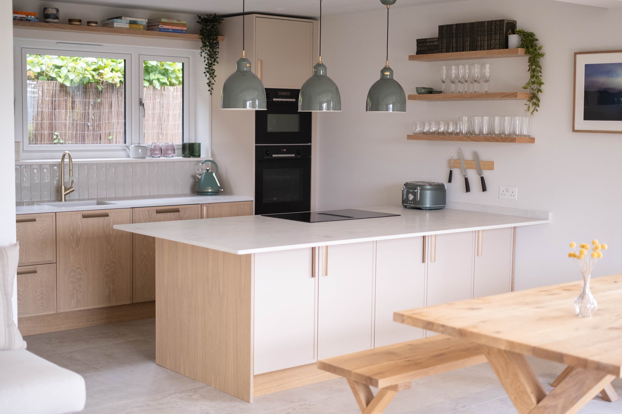 A modern kitchen with light wood cabinets, a white countertop, and a large window with a view of greenery outside. There are three gray pendant lights hanging above the island, and open shelves with books, plants, and glassware on the wall. A toaster, knives, and a green kettle are on the counter, with a small vase of yellow flowers in the foreground.