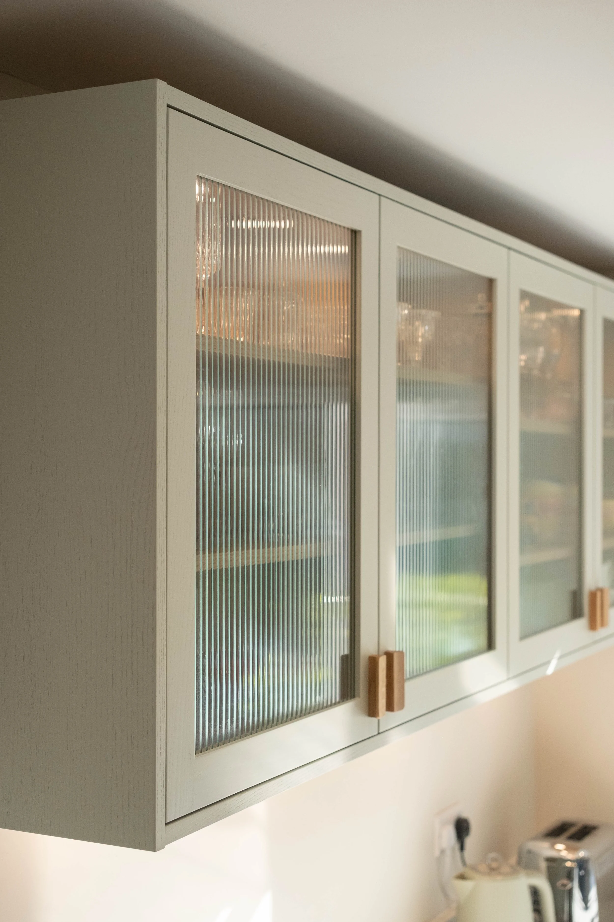 White wall-mounted cabinet with glass doors, wooden handles, and a textured glass surface, in a kitchen with a kettle and toaster on the countertop below.