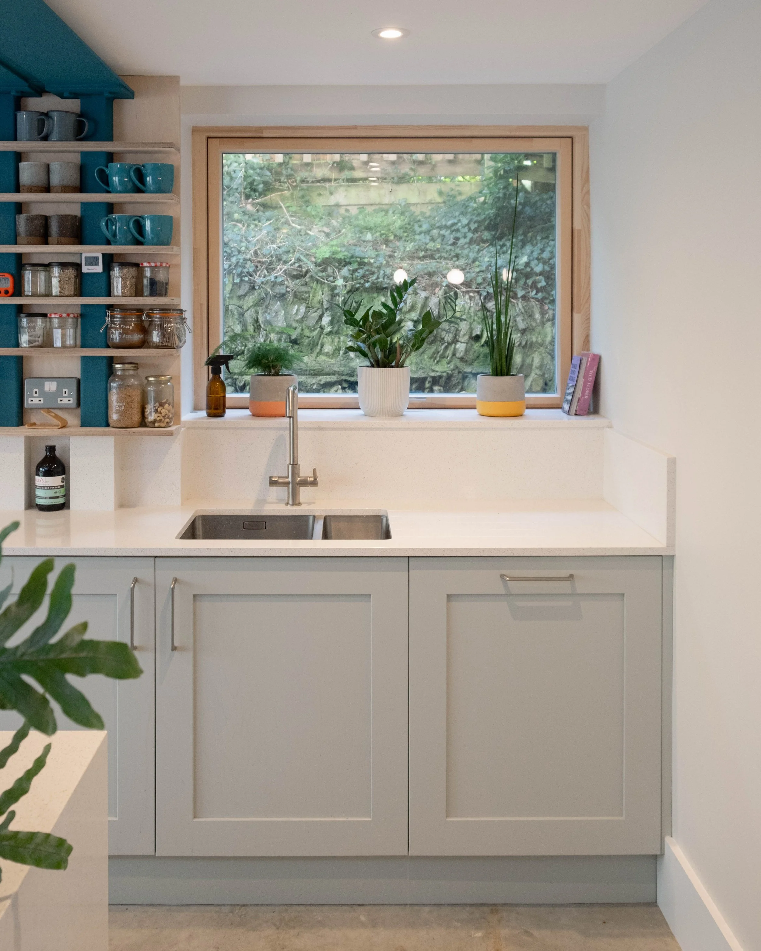 White kitchen sink with a faucet, three potted plants on the windowsill, and a window showing trees outside; open shelves with mugs, jars, and containers on the left side.