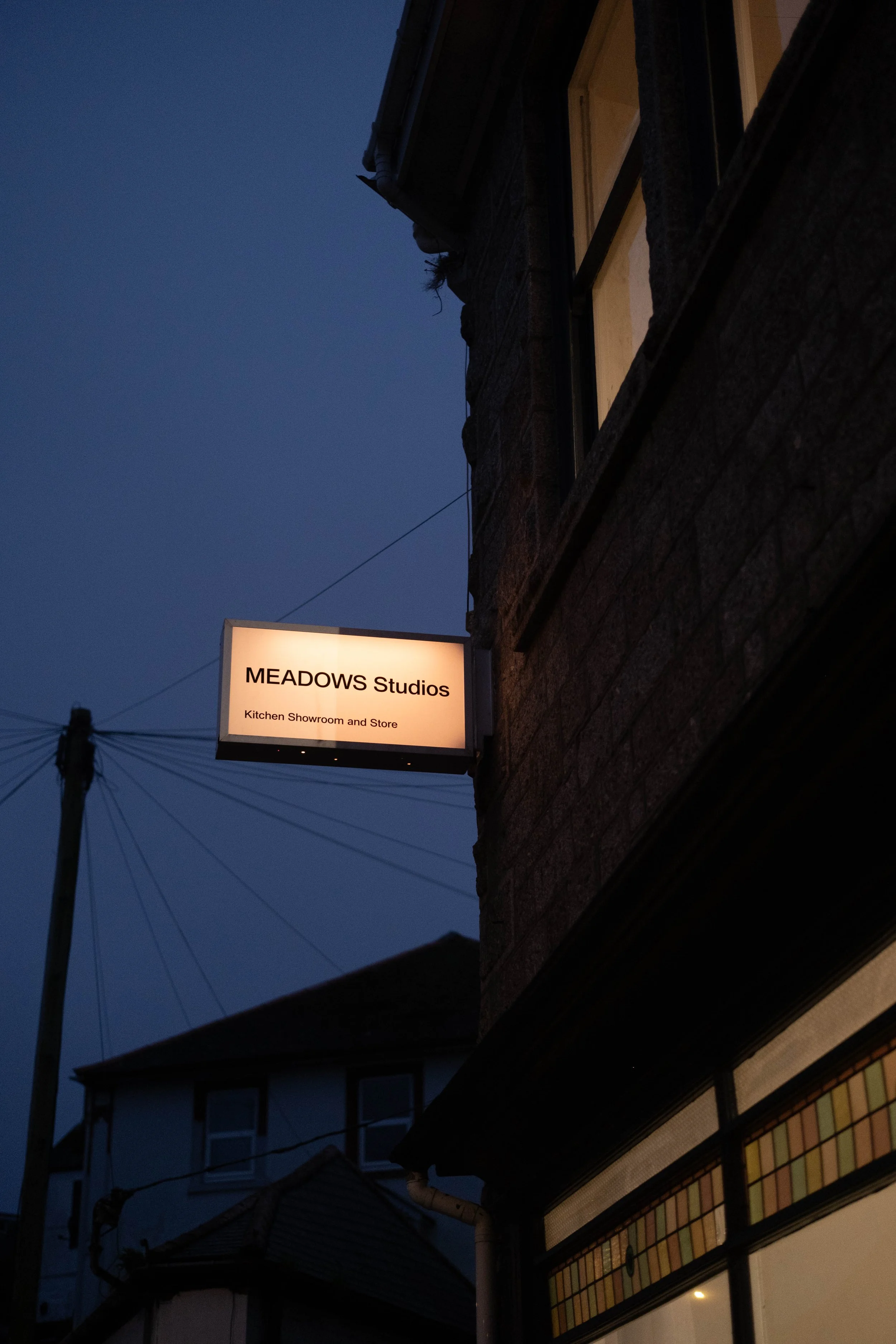 A street scene at dusk showing a sign for MEADOWS Studios, a kitchen showroom and store, attached to a building with a brick exterior. A utility pole with wires is visible in the background, and part of a building with colorful stained glass windows is at the bottom right of the image.