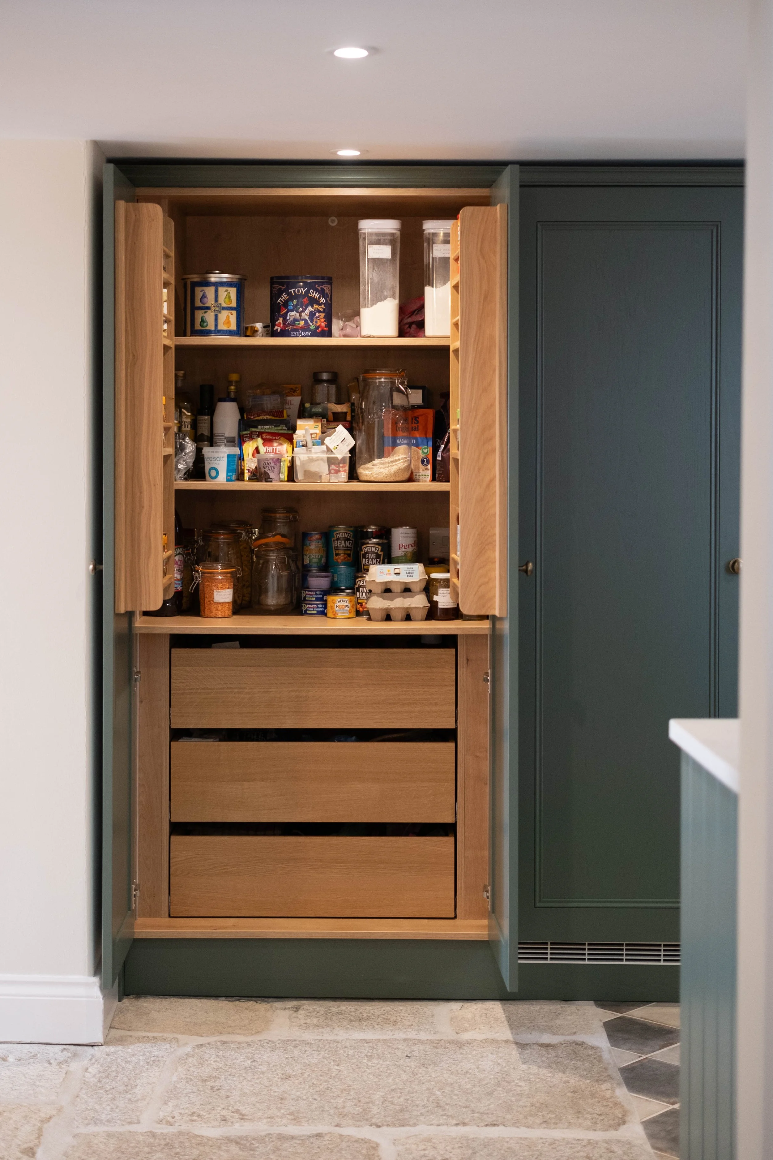 Open kitchen cabinet with wooden shelves containing jars, canned foods, and storage containers, situated on a wall with green trim, adjacent to a white wall and tiled floor.