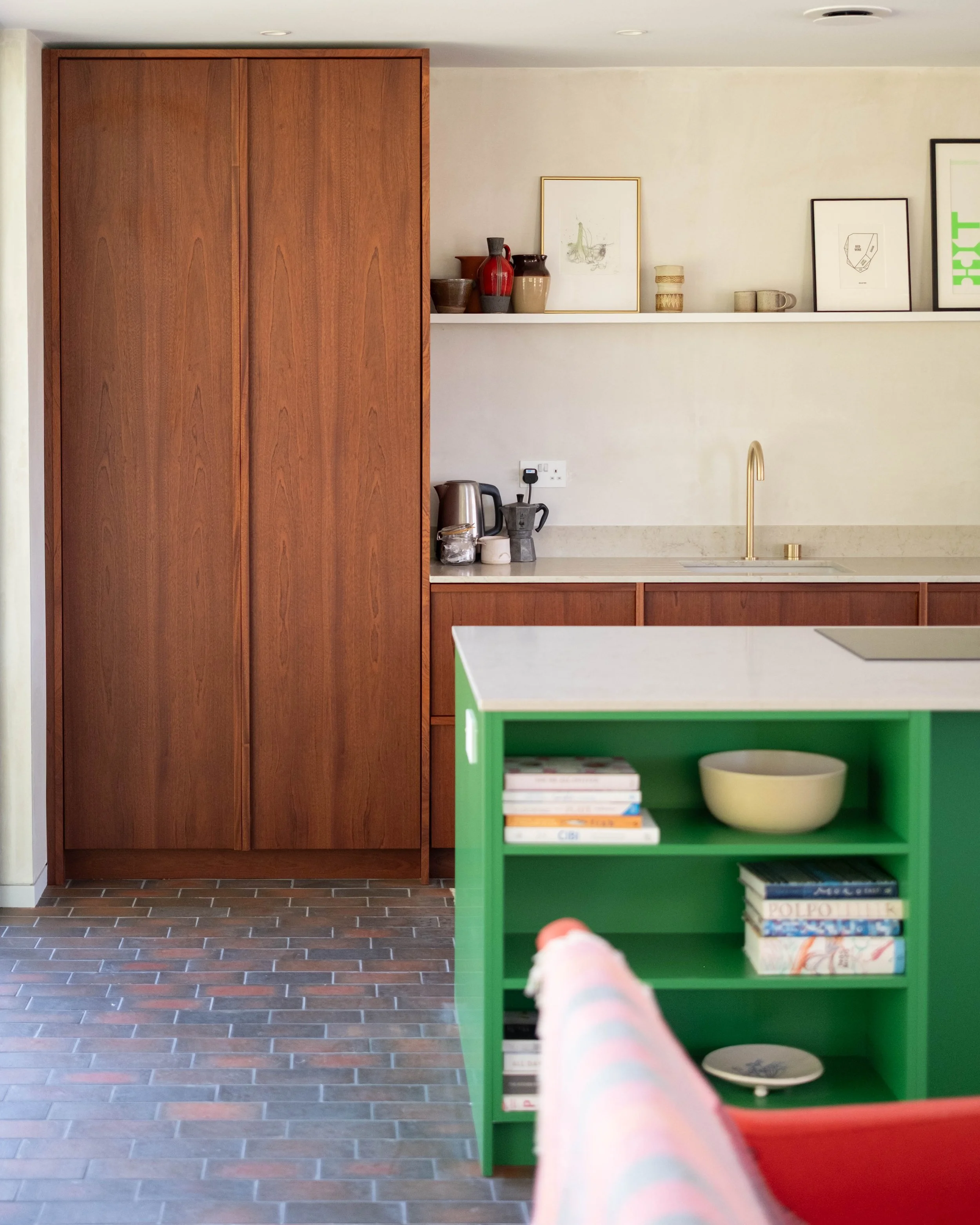 A kitchen with wooden cabinets, a white countertop, and a green open shelf. Items include books, a bowl, a kettle, and decorative vases. The floor is tiled with brown bricks, and part of a person’s leg in pajama pants is visible in the foreground.