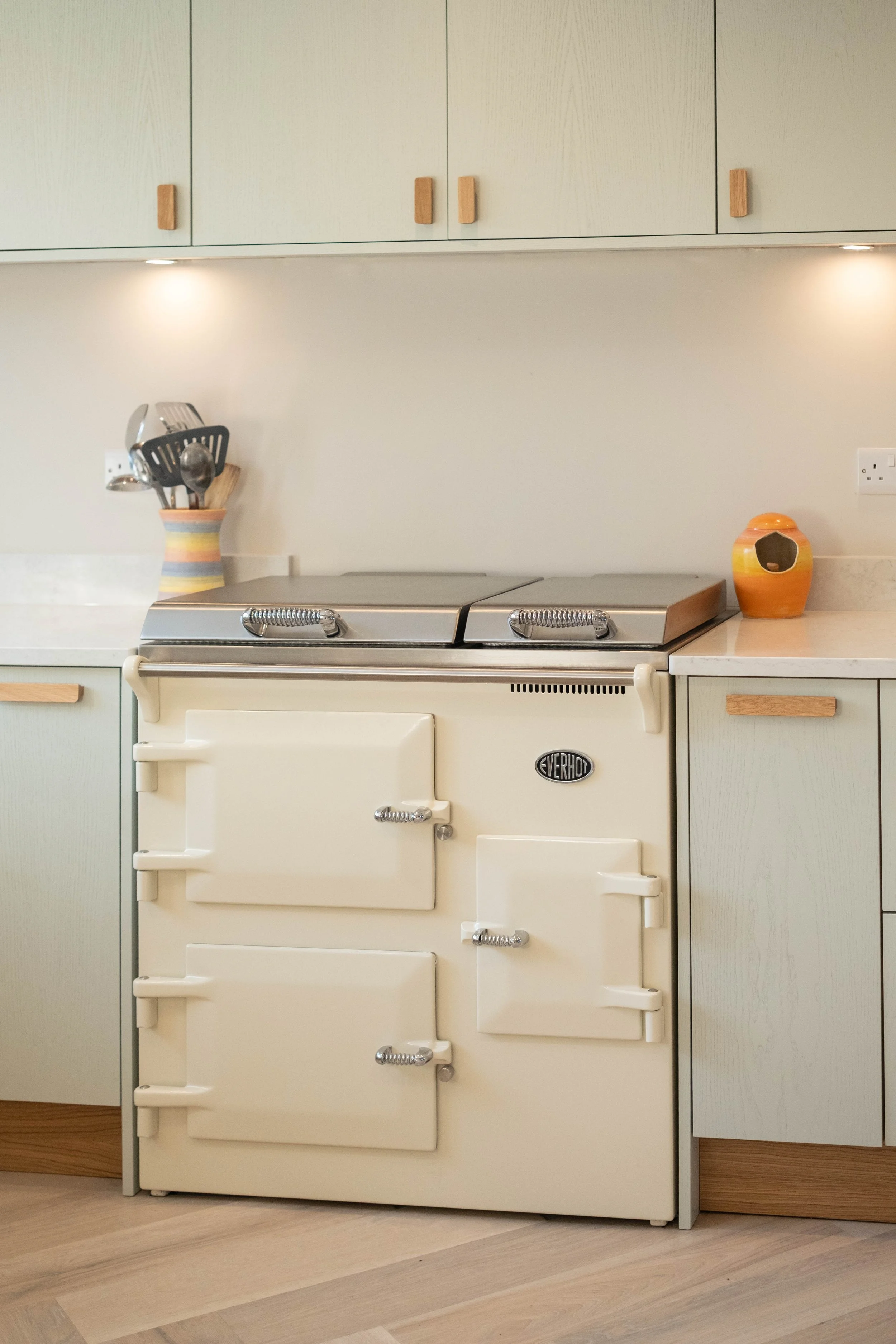A vintage cream-colored electric stove with multiple oven doors and chrome handles, situated in a kitchen with light-colored flooring, cream cabinets, and a countertop with a striped utensil holder and a decorative orange and yellow ceramic jar.