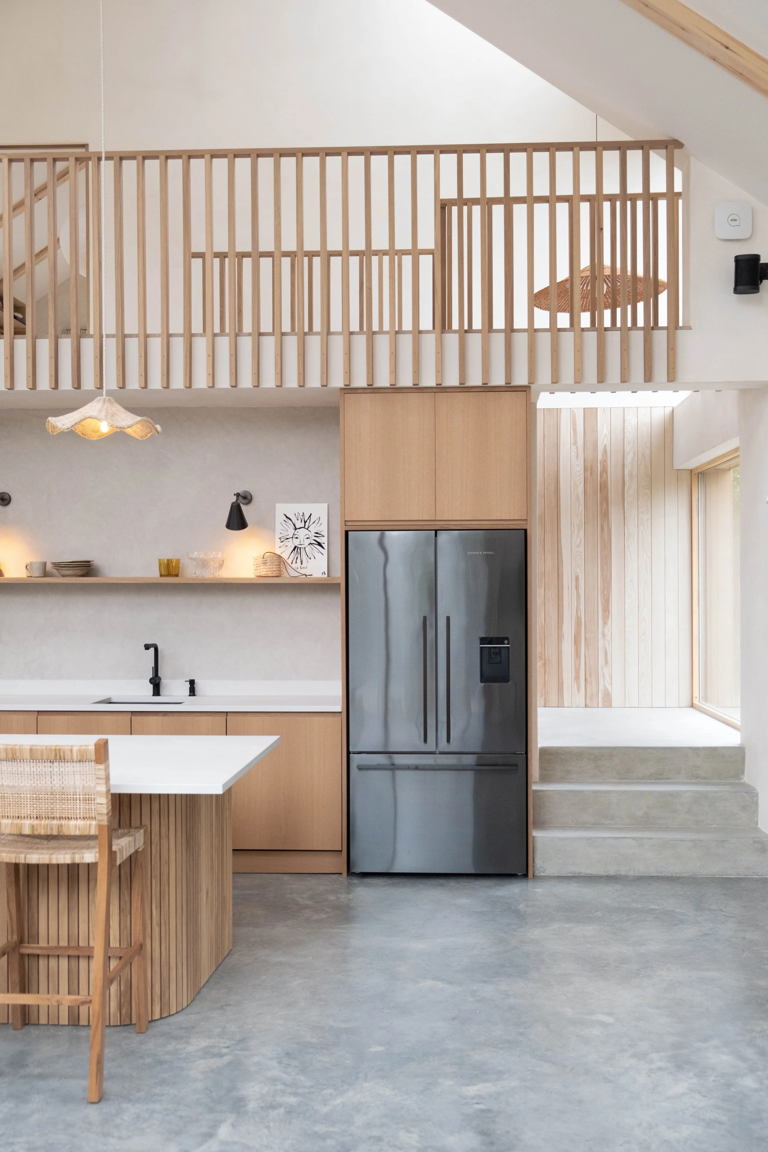 Modern kitchen with light wood cabinets, stainless steel refrigerator, and a wooden dining table with a woven chair.