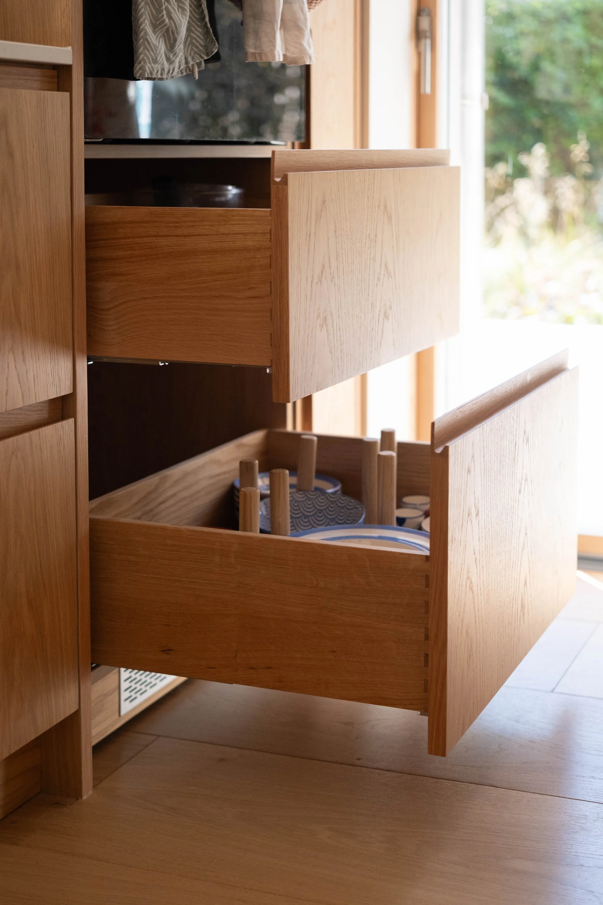 Open wooden kitchen drawer with blue and white patterned bowls inside, and a second drawer above it. The kitchen has wooden cabinetry and a large window with greenery outside.
