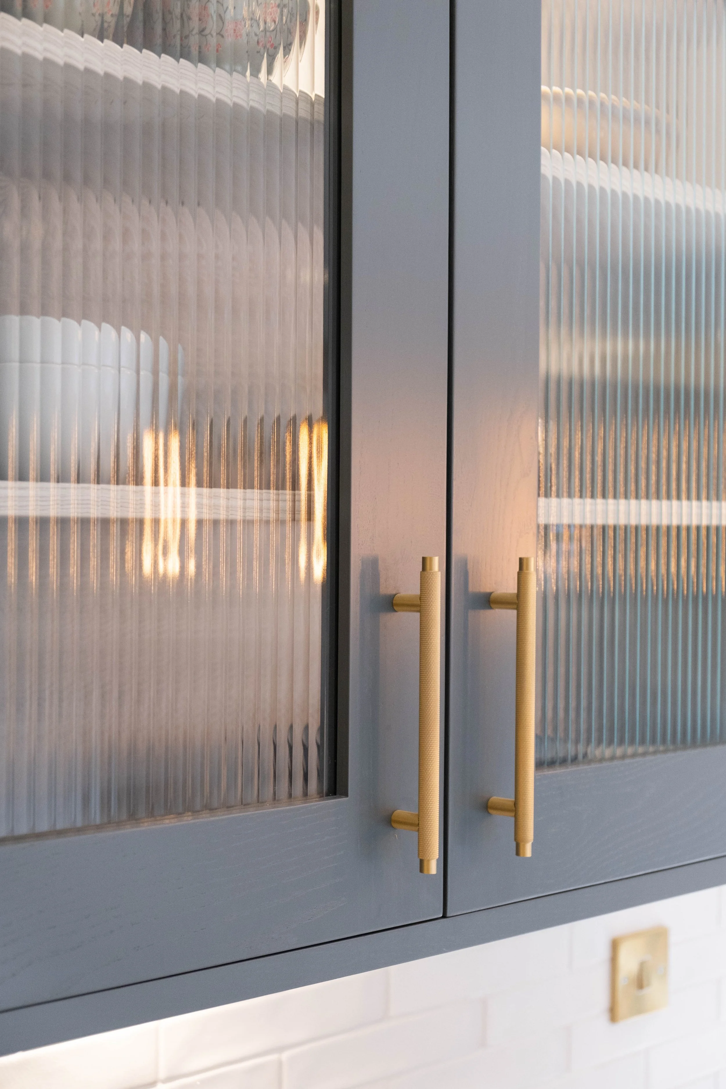 Close-up of kitchen cabinet with textured glass doors and light-colored handles, with a white tiled wall underneath.