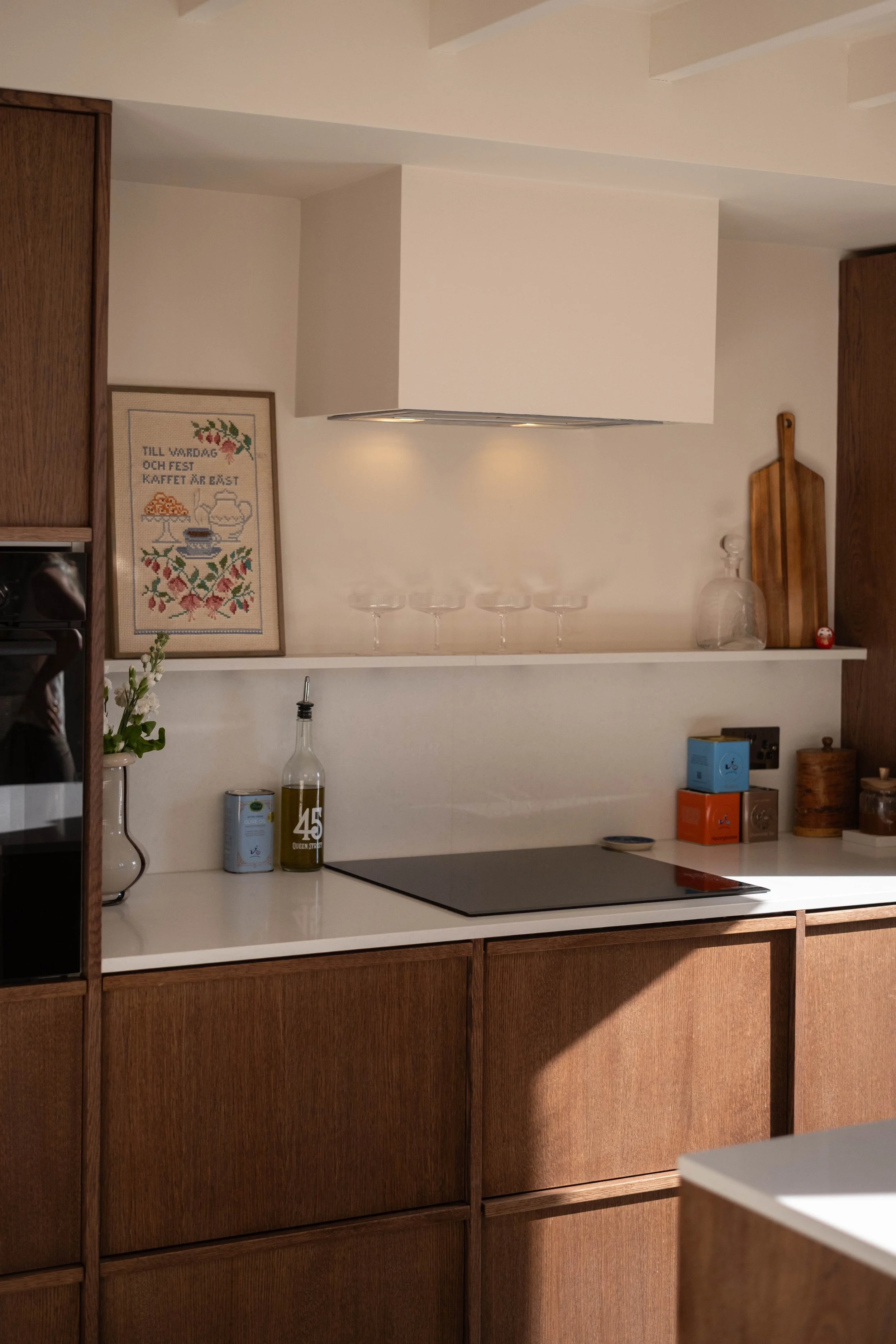 A modern kitchen with white countertops, wooden cabinets, and a white wall. Items on the countertop include glass bottles, canned goods, a wrapped package, and a framed cross-stitch picture that says 'Till vardag och fest kaffet är bäst' with a teapo
