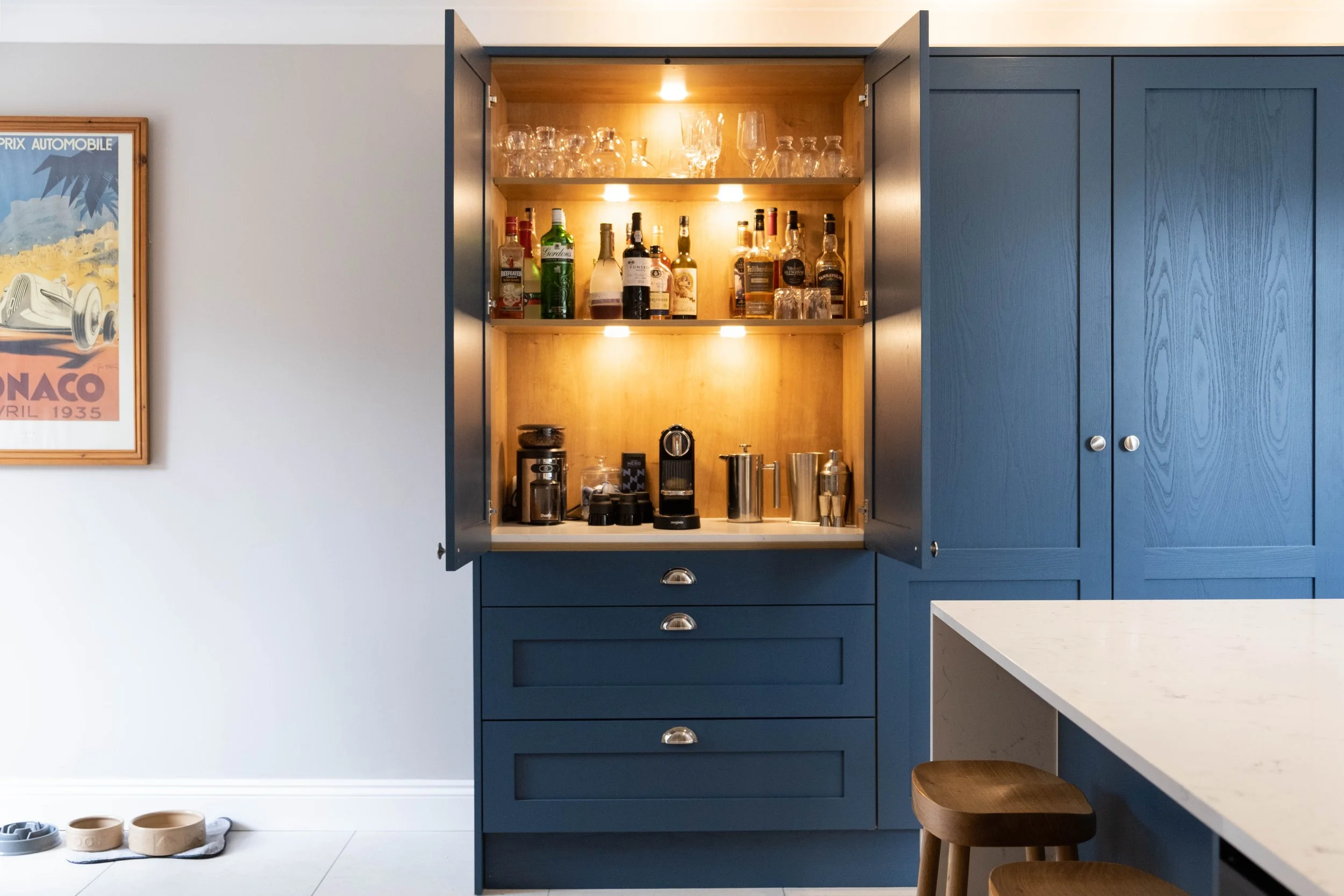 Open blue kitchen cabinet with liquor bottles, glasses, and coffee station inside, next to a light-colored wall with a framed poster, a white countertop, and wooden stools.