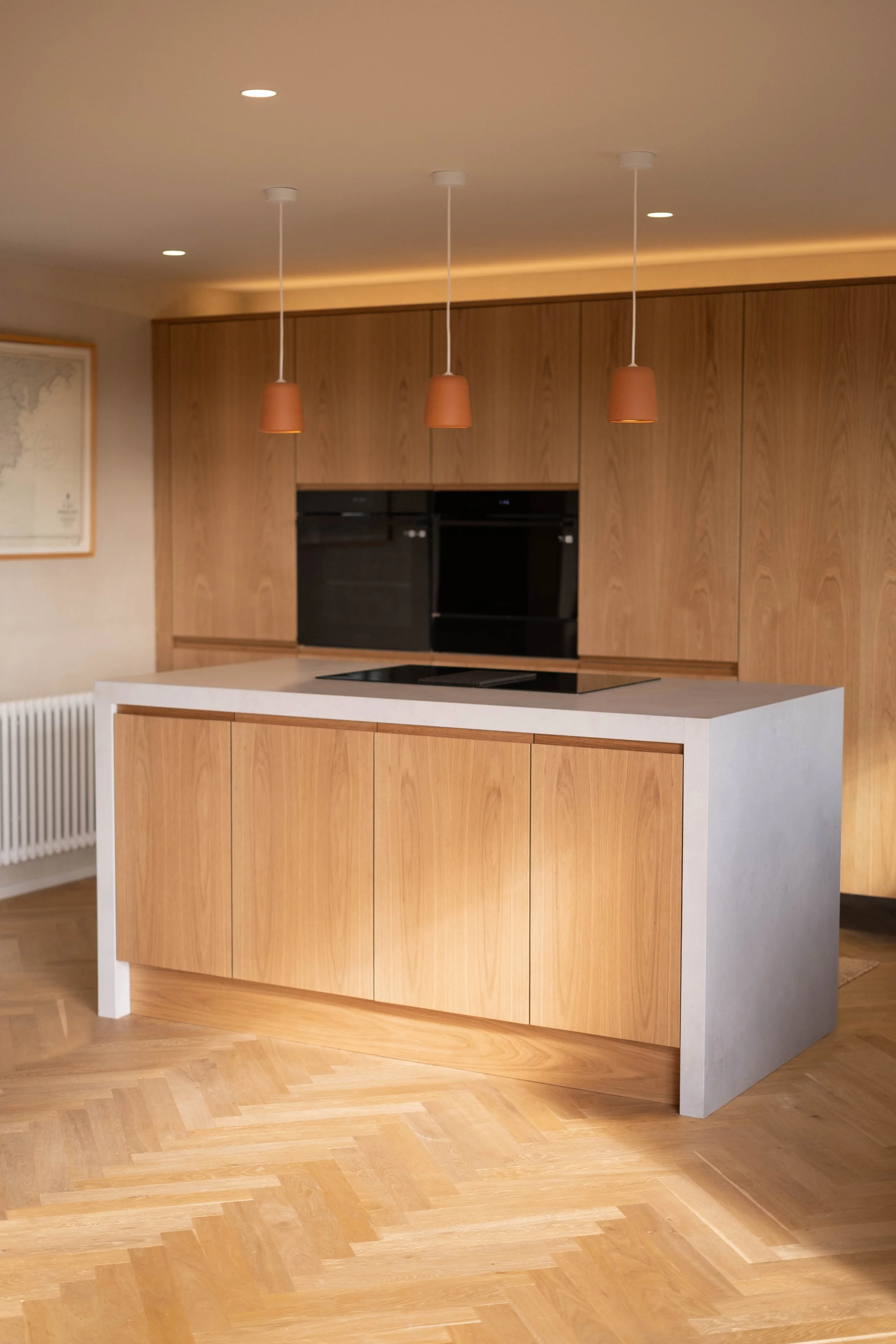 Modern kitchen with wooden cabinets, black built-in oven, white island with wooden doors, floating terracotta pendant lights, and a parquet wood floor.