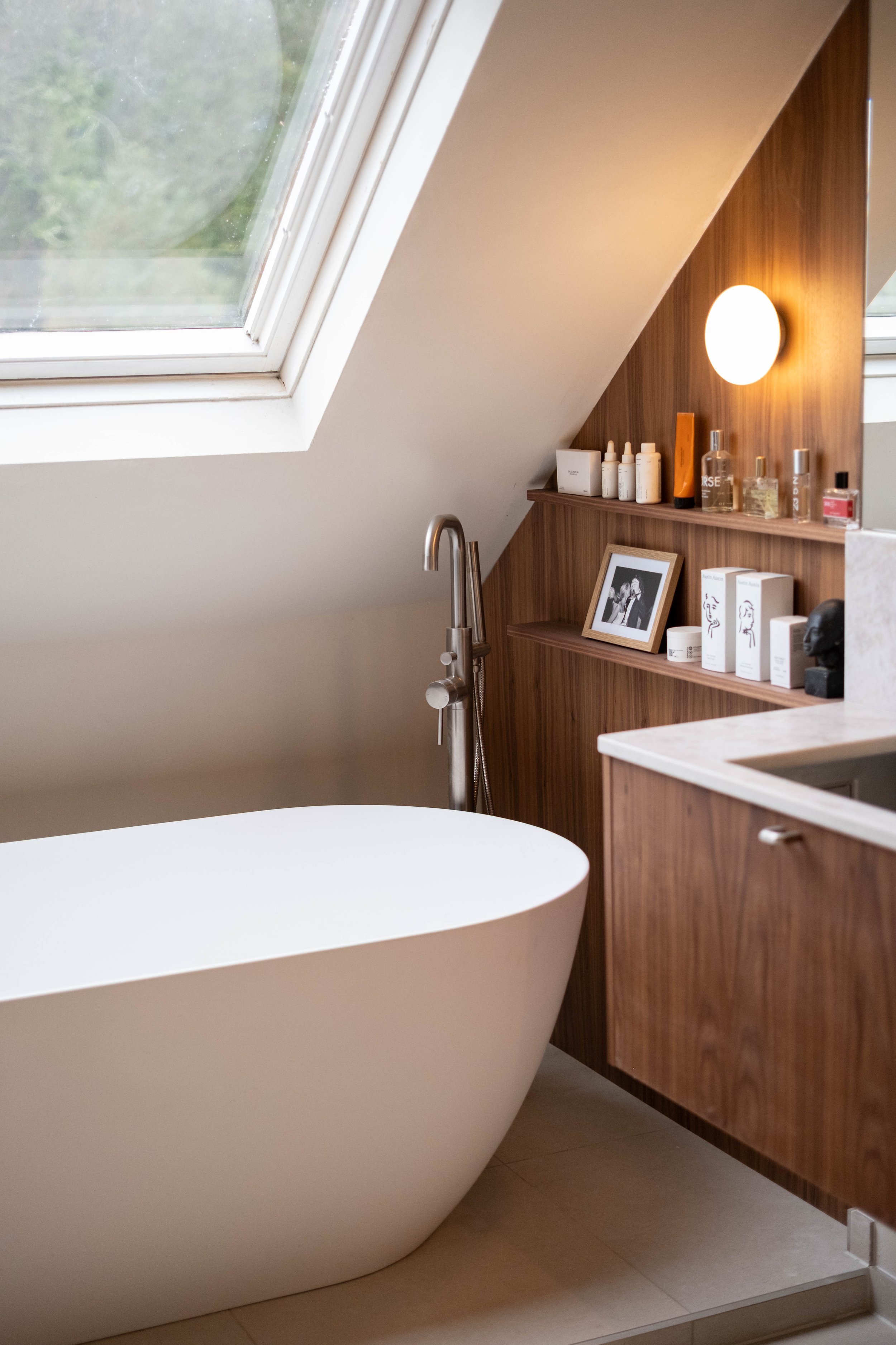 A modern bathroom with a freestanding bathtub, skylight window, and wooden vanity with various skincare products and a framed black-and-white photograph.