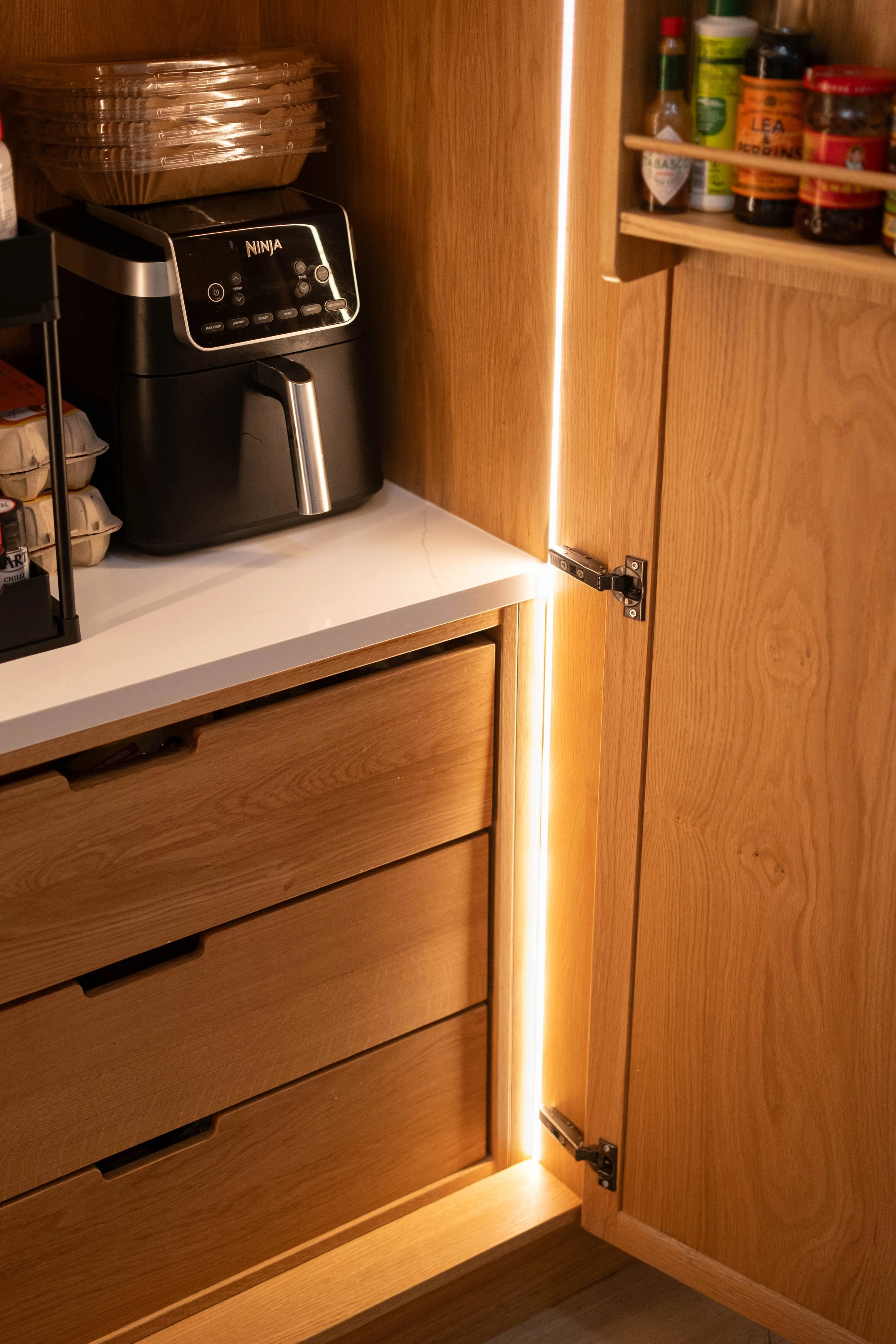 Open wooden kitchen cabinet with a concealed LED light strip on the side, showing a kitchen countertop with spices and cooking essentials.