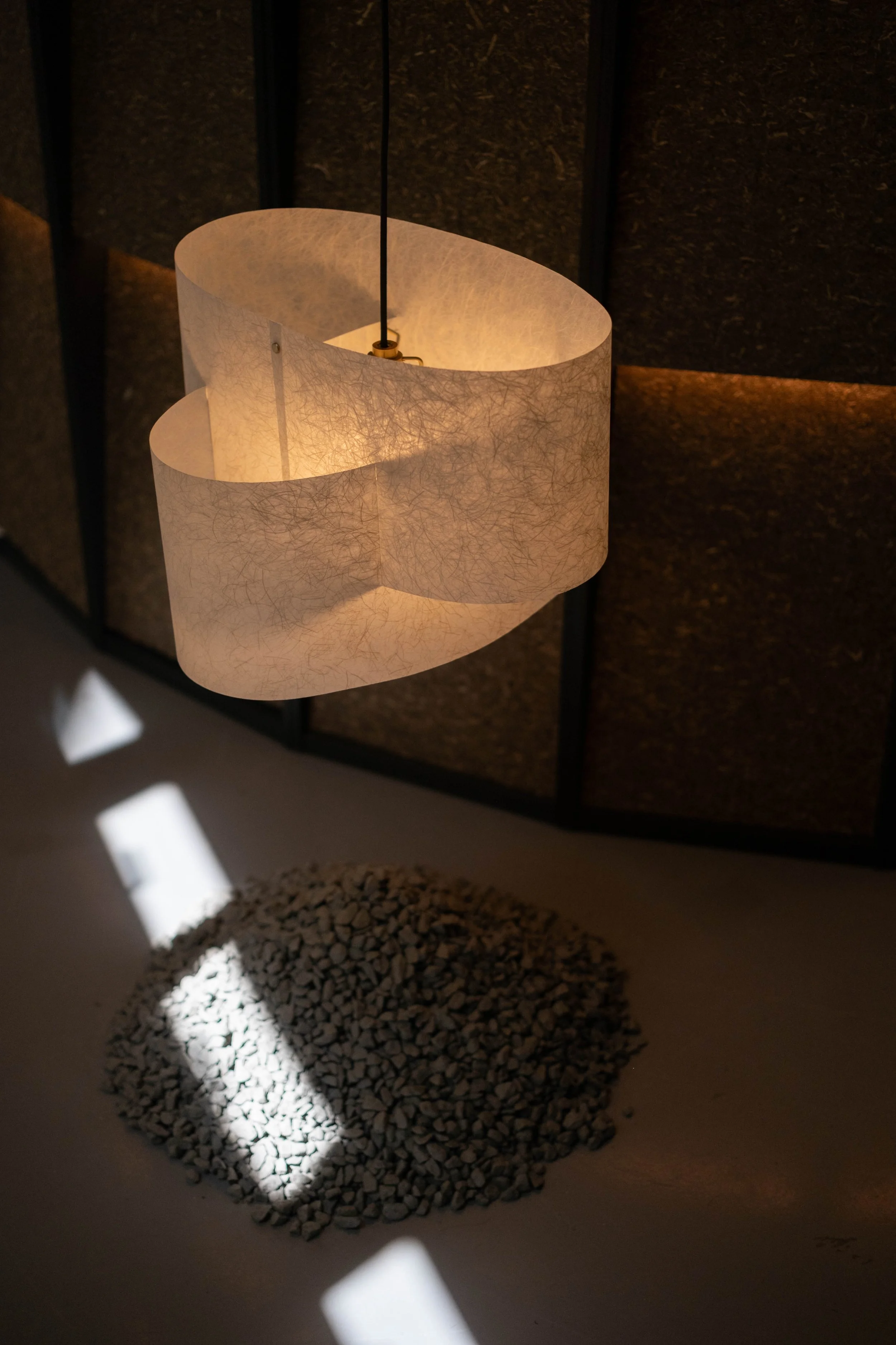 A modern pendant lamp with a textured white paper shade hangs above a circular pile of small white rocks on the floor. Shadows and light cast artistic patterns on the floor.