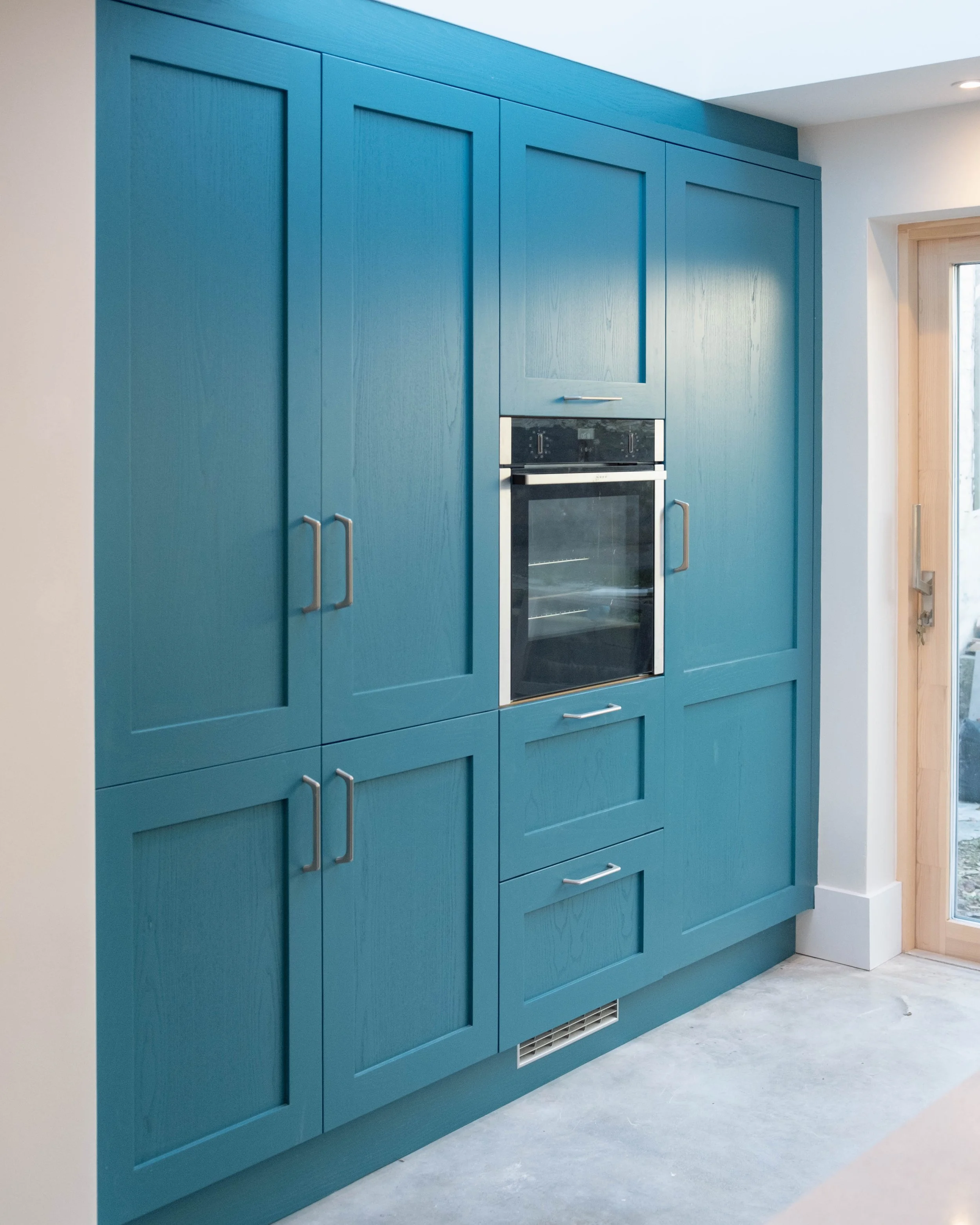 Blue kitchen cabinets with silver handles, built-in oven, white walls, and a window to the right, in a modern kitchen.