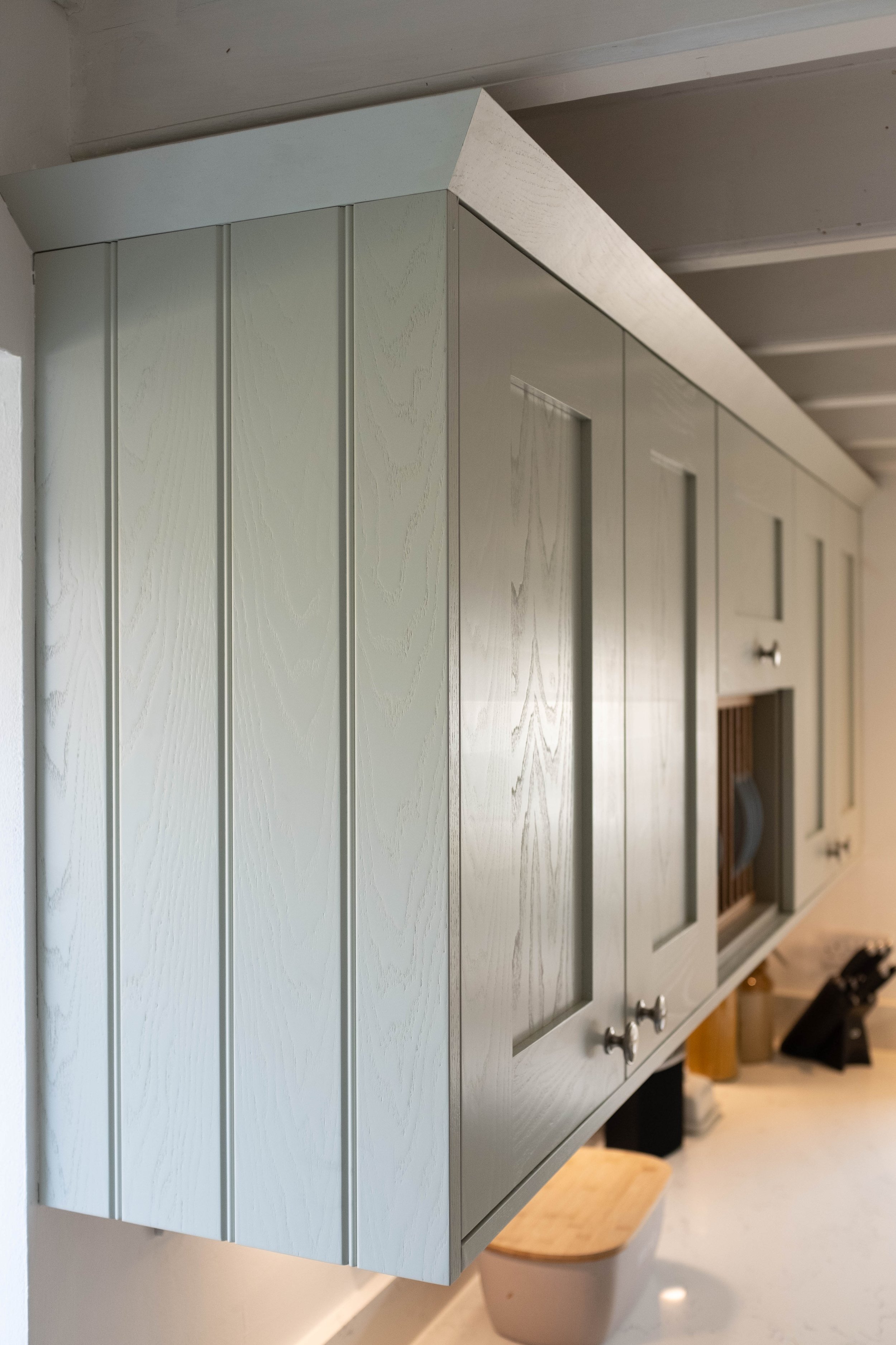 Light gray kitchen cabinet with vertical panels and round silver handles, seen from an angle in a kitchen.